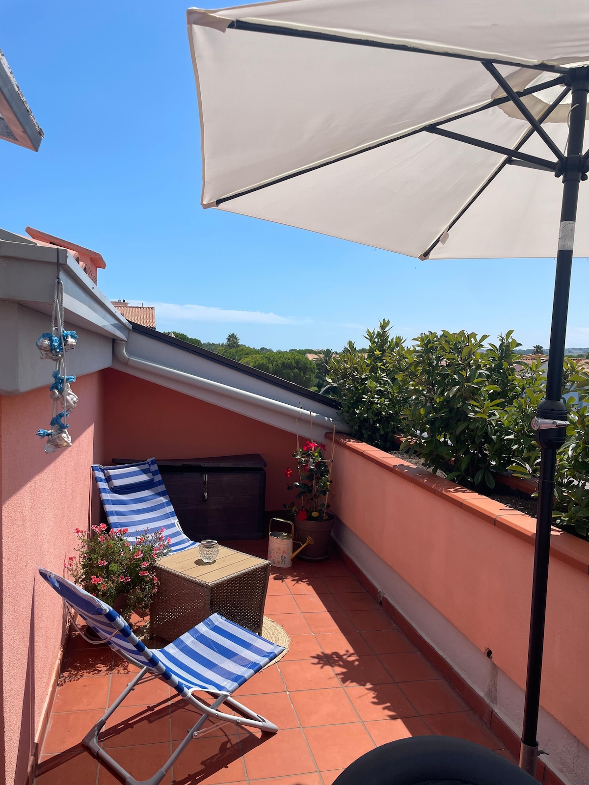 A sunlit terrace features two striped lounge chairs under a large umbrella. Potted plants and flowers add greenery to the vibrant pink walls, creating a relaxing outdoor space. A small table is positioned between the chairs, inviting guests to enjoy the view.