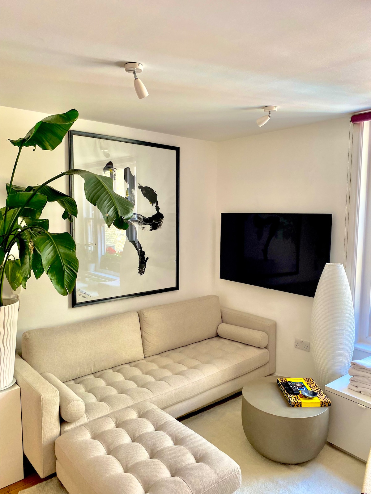 A modern living area is presented, featuring a light-colored sectional sofa and a round, metallic table. A large framed artwork is displayed on the wall, accompanied by a tall potted plant. Ambient light is provided by recessed ceiling fixtures and natural light from nearby windows.