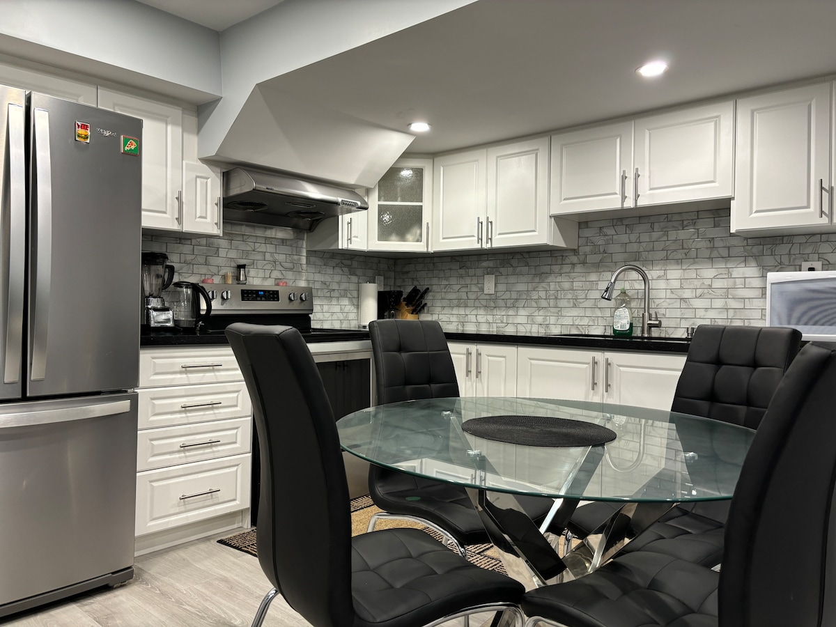 A modern kitchen features white cabinetry, sleek stainless steel appliances, and a marble backsplash. A glass dining table with black chairs is positioned centrally, offering a contemporary dining area illuminated by overhead lights.