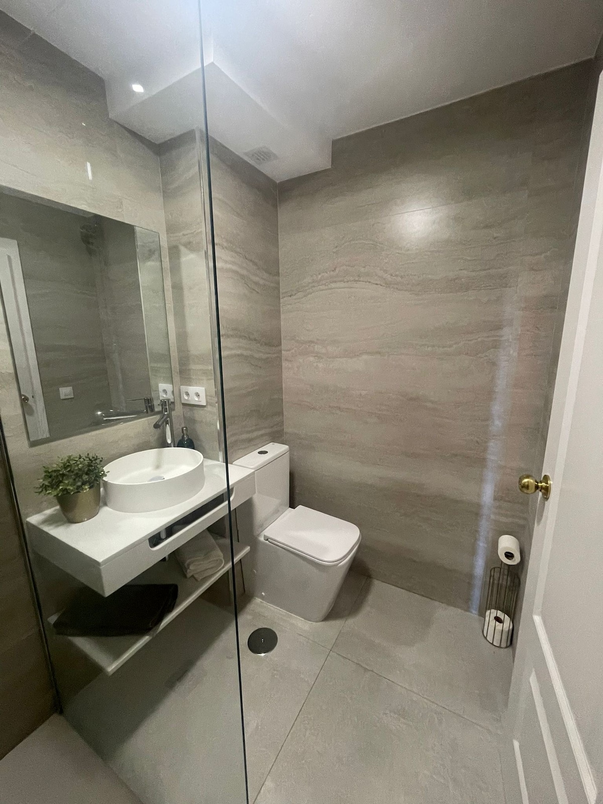 A modern bathroom features a glass shower enclosure, a wall-mounted sink, and a sleek toilet. Natural stone tiles create a soothing ambiance, while a small plant adds a touch of greenery. Shelving holds neatly arranged toiletries, enhancing the contemporary design.