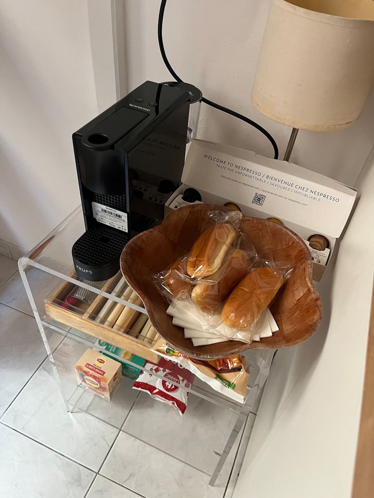A coffee machine is displayed next to a transparent acrylic stand. A woven bowl contains packaged bread rolls, while a neatly arranged selection of coffee pods is visible below. Light reflects off the surfaces, offering a clean and modern look.