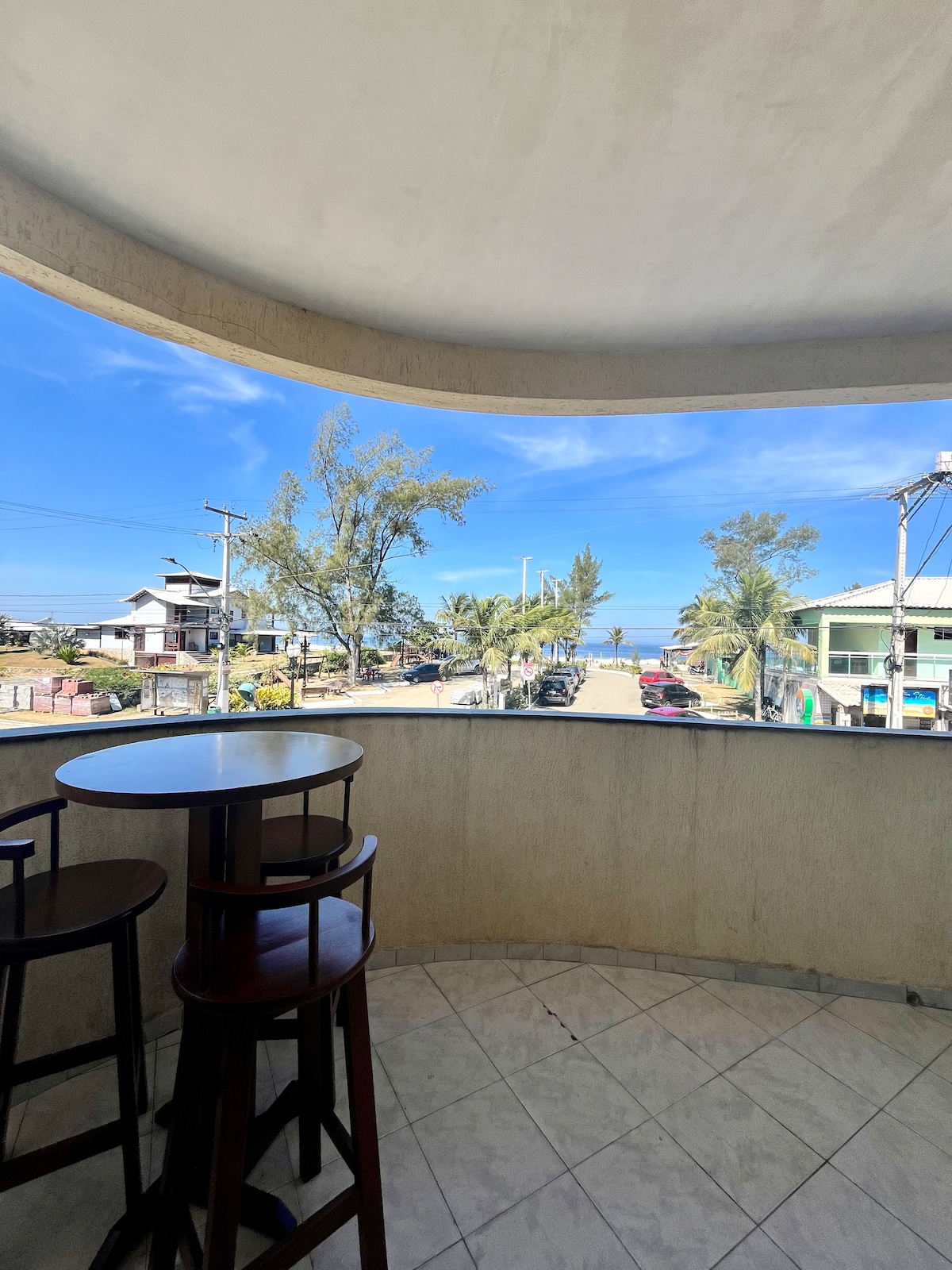 A spacious balcony features two high stools at a circular table, offering an unobstructed view of the street lined with palm trees. The sea is visible in the distance, with a clear blue sky enhancing the scene.