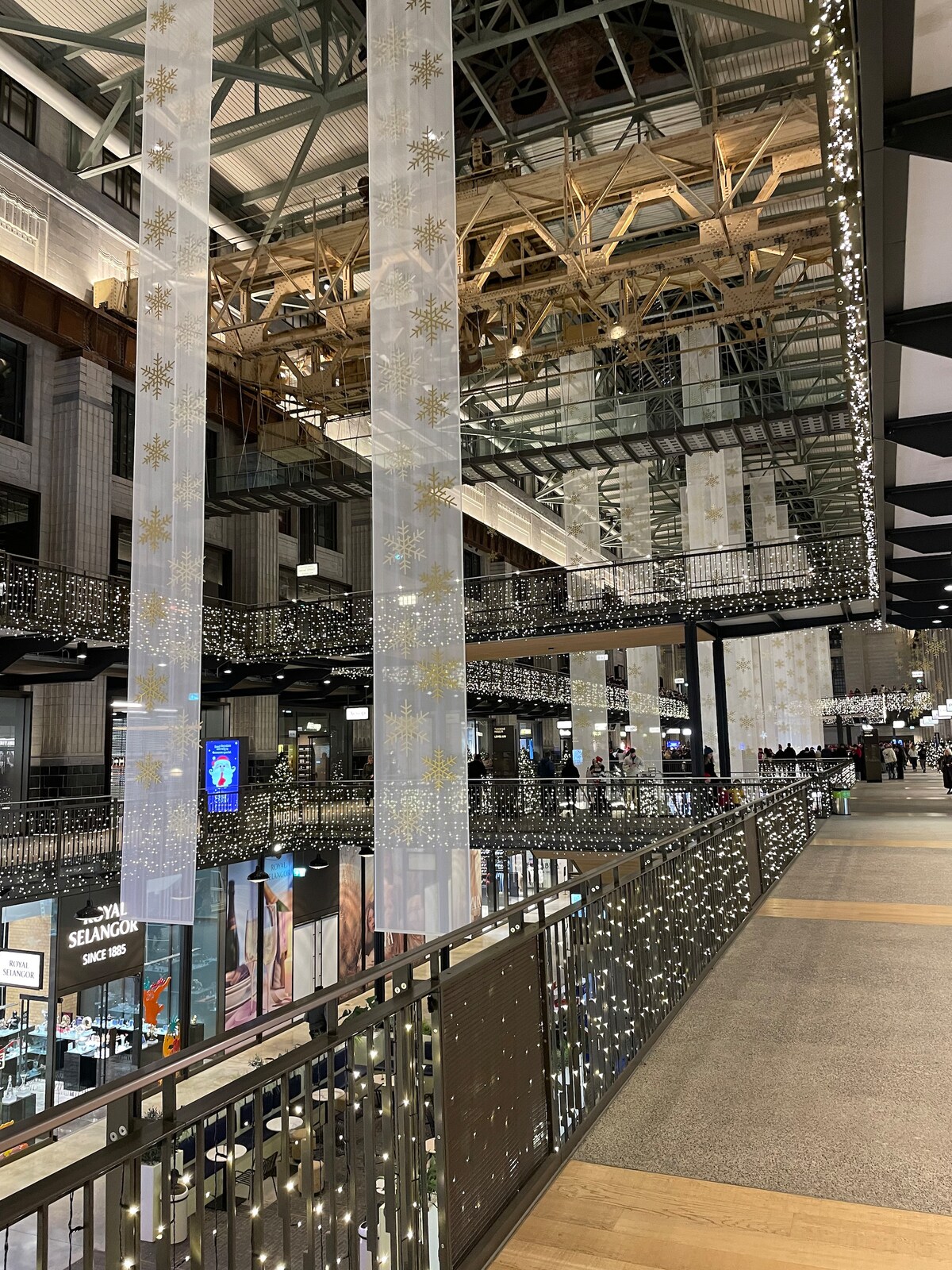 The interior of the Battersea Power Station development is illuminated by numerous lights, creating a vibrant atmosphere. Tall ceilings and wooden structures are visible, along with multiple levels connected by open railings. Retail spaces can be seen below, bustling with visitors.