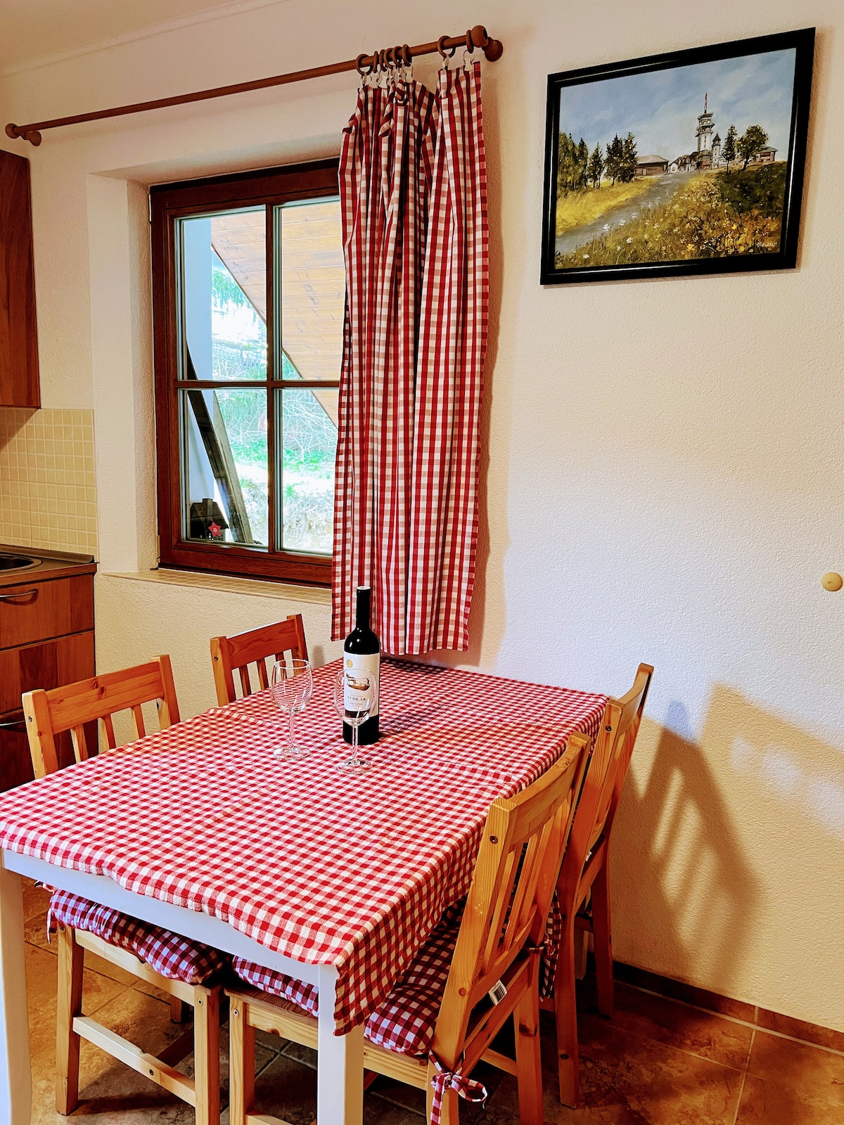 A dining area features a white table covered with a red and white checkered tablecloth, surrounded by four wooden chairs. A bottle of wine and two glasses are placed at the center of the table. A framed landscape painting hangs above the window, where red gingham curtains are drawn.