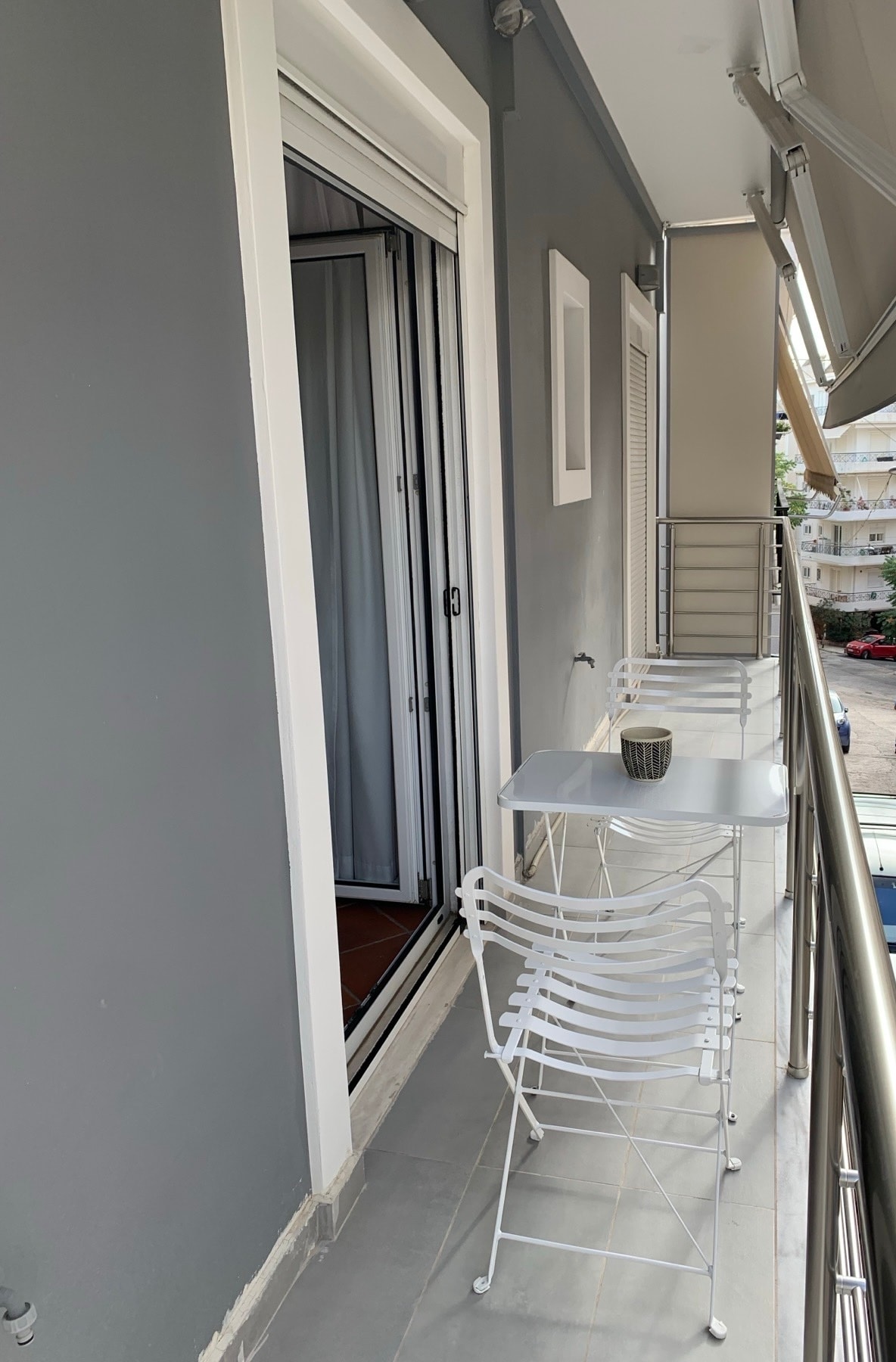 A narrow balcony is featured, equipped with a small white table and two folding chairs. The sliding door leads into the apartment, while the gray walls provide a modern backdrop. Sunlight enhances the space, and views of the surrounding area are visible beyond the railing.