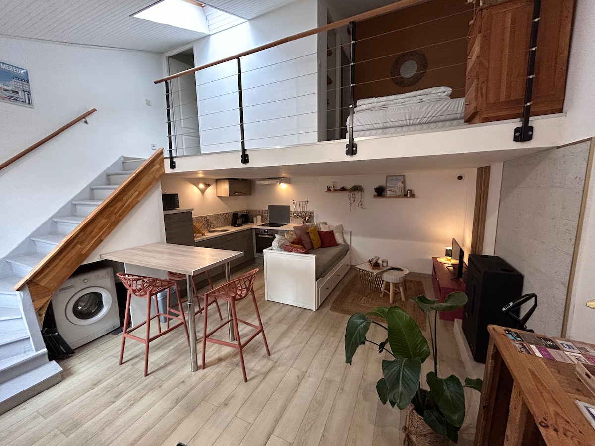The open living area features a double banquette bed positioned next to a modern kitchenette. A dining table with four chairs is situated nearby. A staircase leads to a mezzanine containing a double bed. Natural light enters through a skylight above.