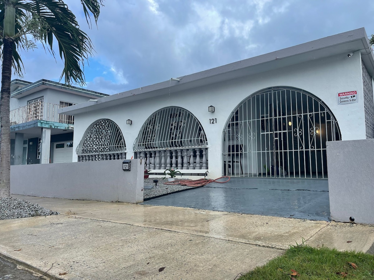 The front view of a single-story house is presented, featuring three large arched windows secured with decorative metal grilles. A smooth concrete driveway leads to a covered entrance, with a well-maintained yard and palm trees flanking the property.