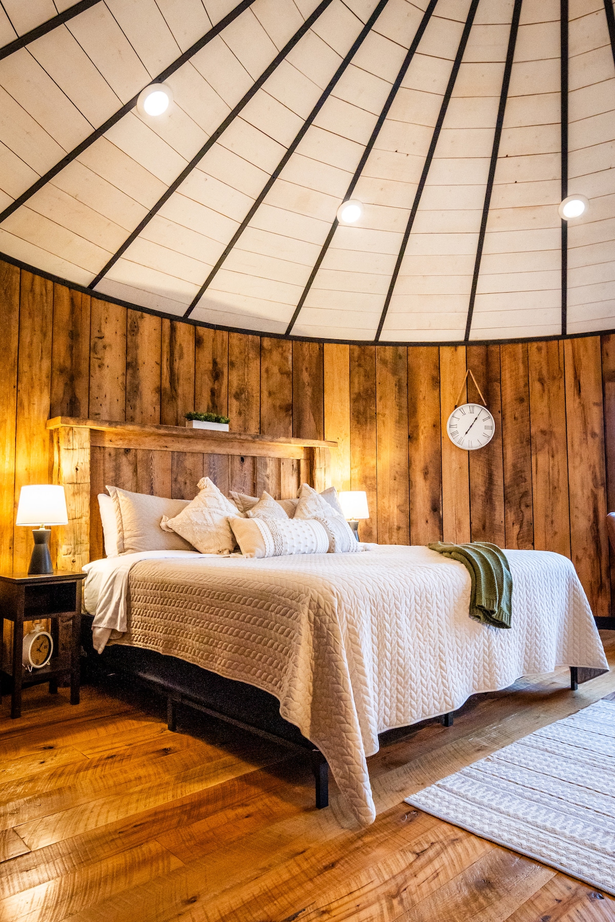 A circular bedroom showcases a spacious bed adorned with white bedding. The wooden walls create a warm environment, while two bedside lamps offer soft lighting. A clock is mounted above the bed, and a textured area rug lies on the hardwood floor.