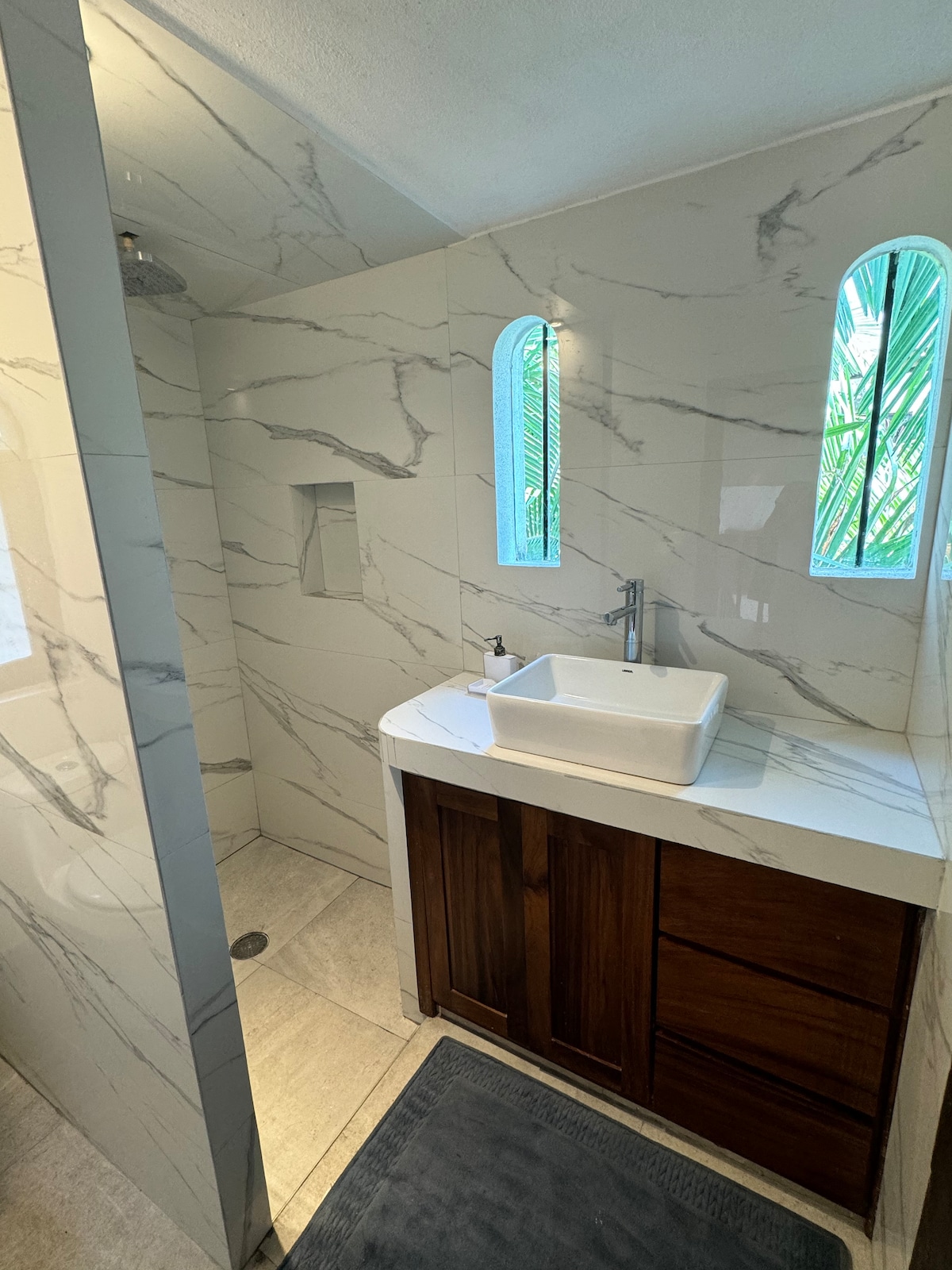 A modern bathroom features textured walls with marble-like tiles and a stylish wooden vanity. A rectangular sink is positioned on the counter, while a glass-enclosed shower area is visible. Natural light filters through small, arched windows surrounded by greenery.