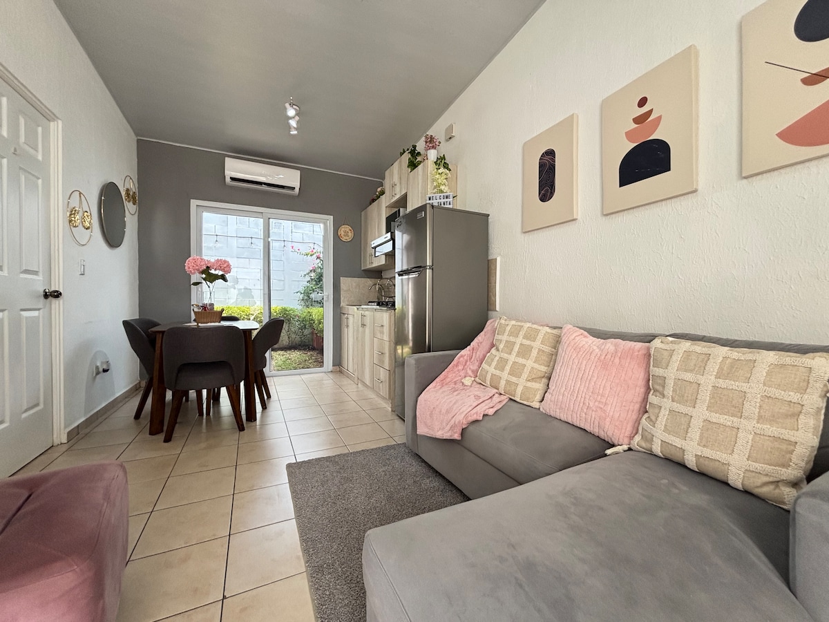 A cozy living area features a light grey sofa with patterned cushions, complemented by a plush rug. A dining table with four chairs is visible, along with an open kitchen showcasing modern appliances. Natural light enters through a large window, enhancing the space.