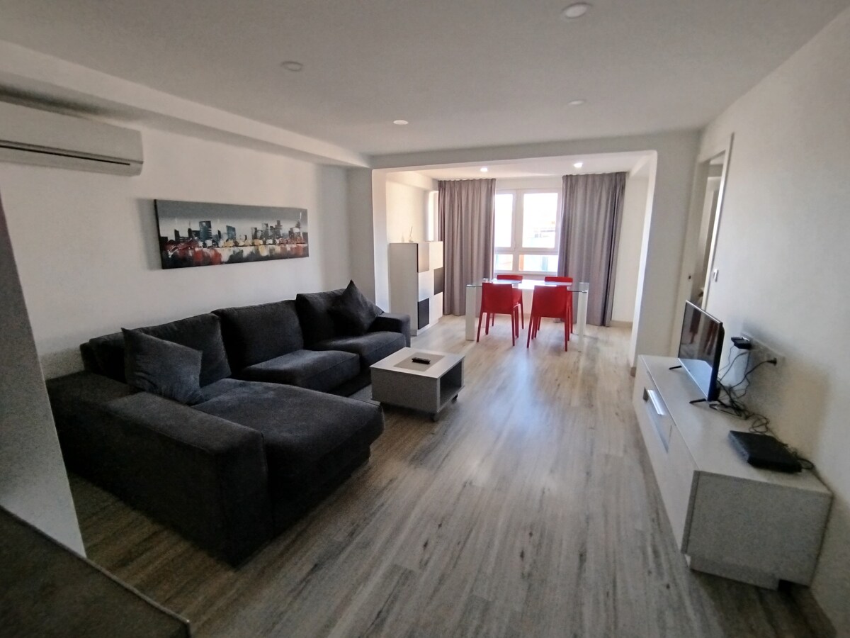 A spacious living area is featured with a large, dark gray sectional sofa and a light-colored coffee table. A flat-screen television is placed on a modern media console. Dining is possible at a small table with red chairs, positioned near a window dressed with light gray curtains.