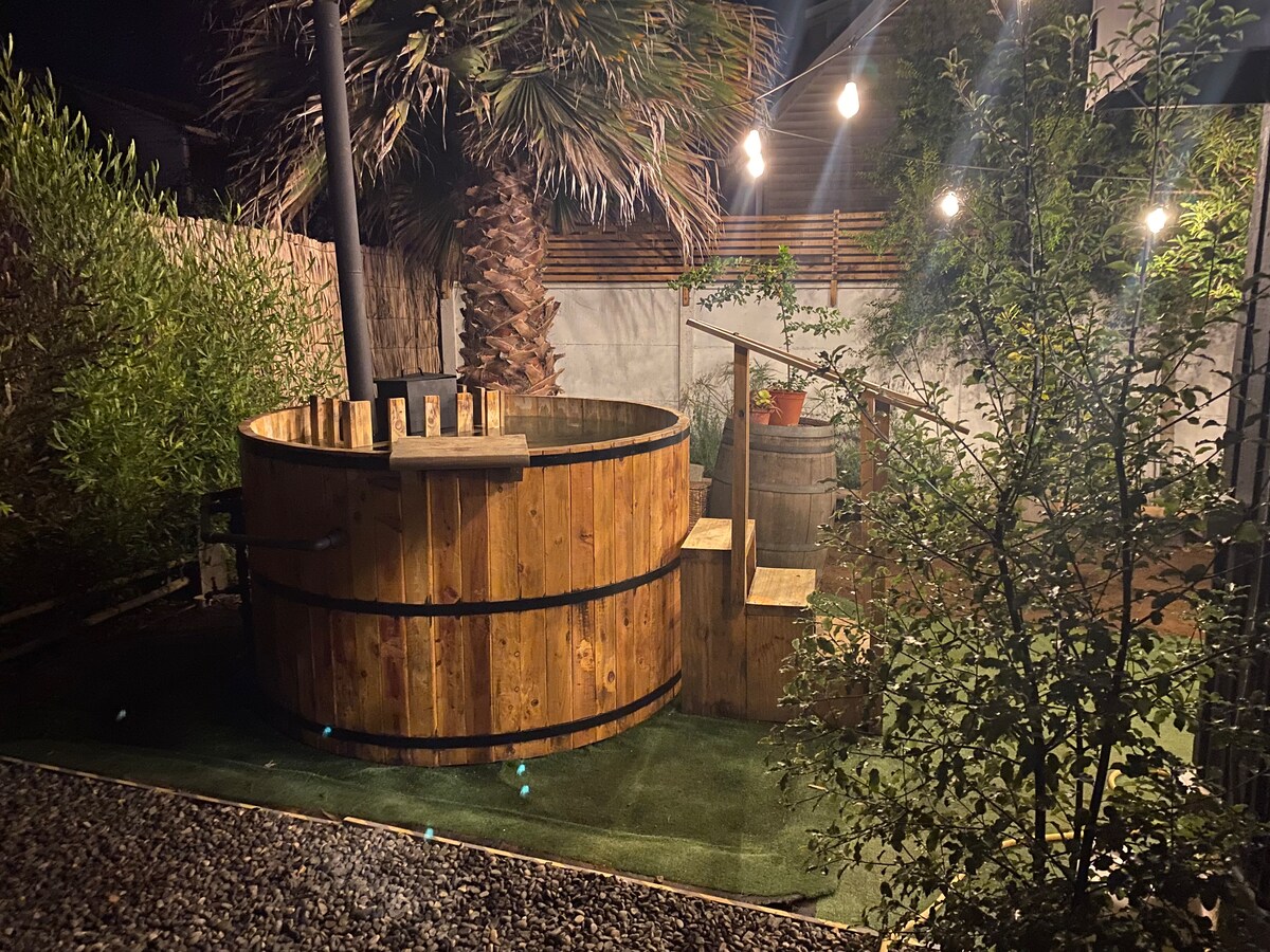 A wooden hot tub is seen surrounded by lush greenery and palm trees. Soft lighting from hanging bulbs illuminates the area, creating a cozy ambiance. A small staircase provides access to the tub, with a barrel nearby enhancing the rustic charm of the outdoor space.