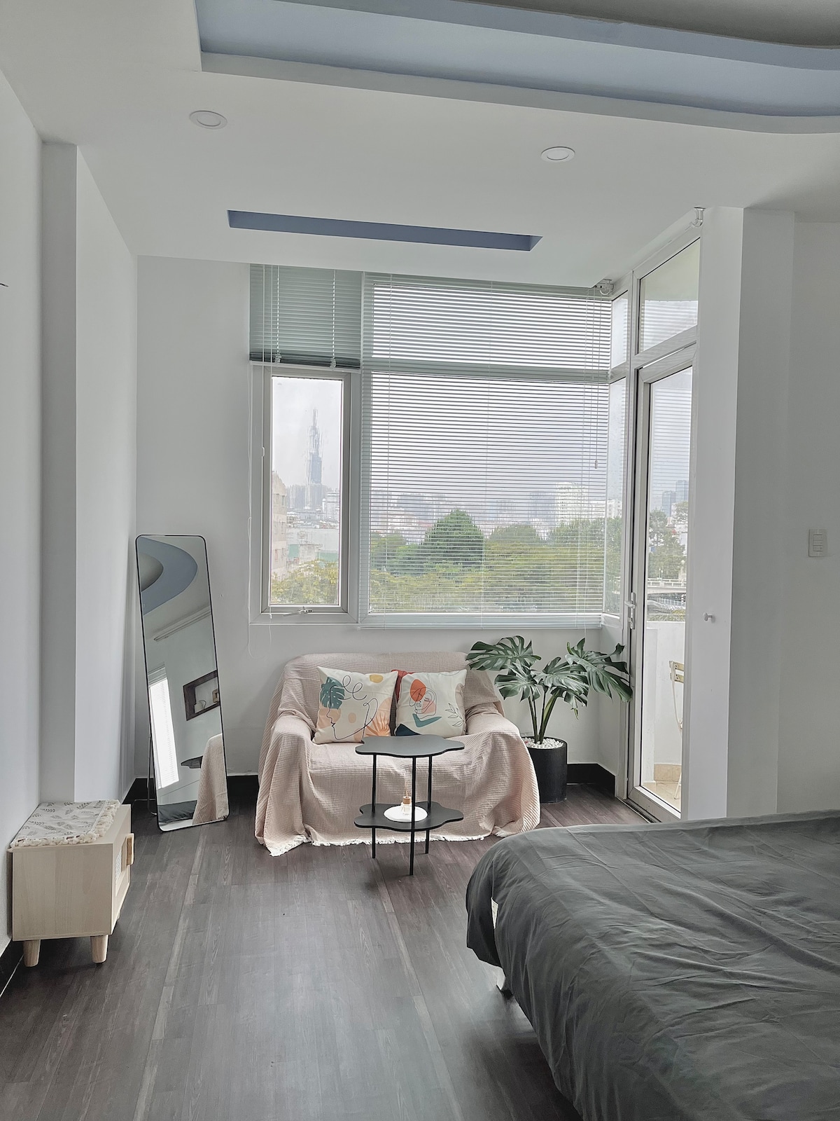 A bright seating area is positioned by large windows, offering views of the city. A cozy chair is adorned with decorative cushions, complemented by a small side table. Soft greenery from nearby plants enhances the inviting atmosphere of the space.