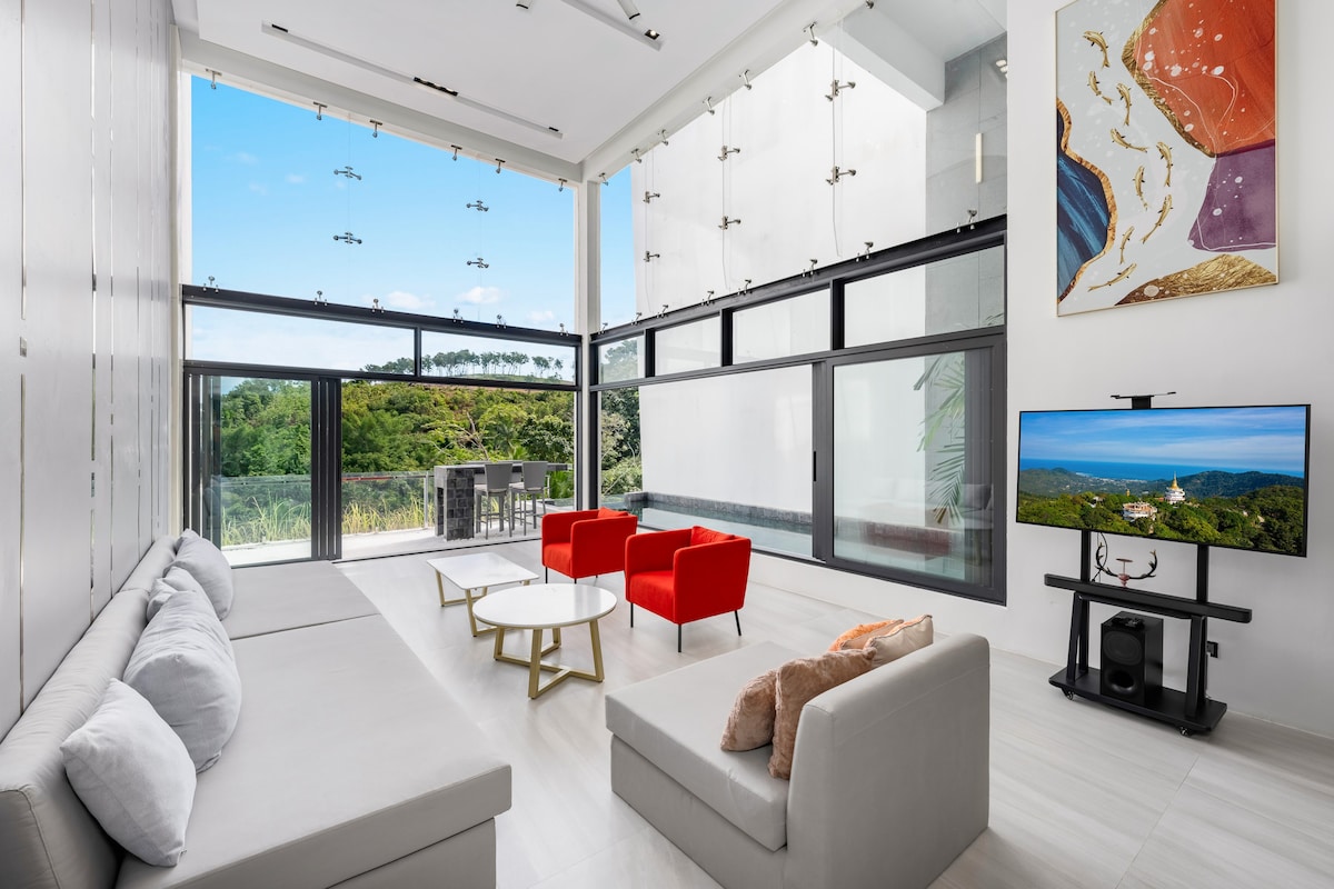 A spacious living area is bathed in natural light, featuring a large sectional sofa and two red accent chairs. A modern coffee table sits in the center, while a wall-mounted TV is positioned near a large window showcasing scenic views.