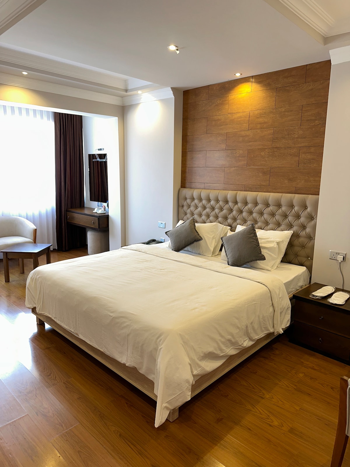 A well-appointed bedroom features a spacious bed with a tufted headboard and neatly arranged pillows. Natural light filters through a large window covered by sheer curtains, illuminating the wooden floor. A small desk and chair are positioned beside the bed, with a light fixture above.