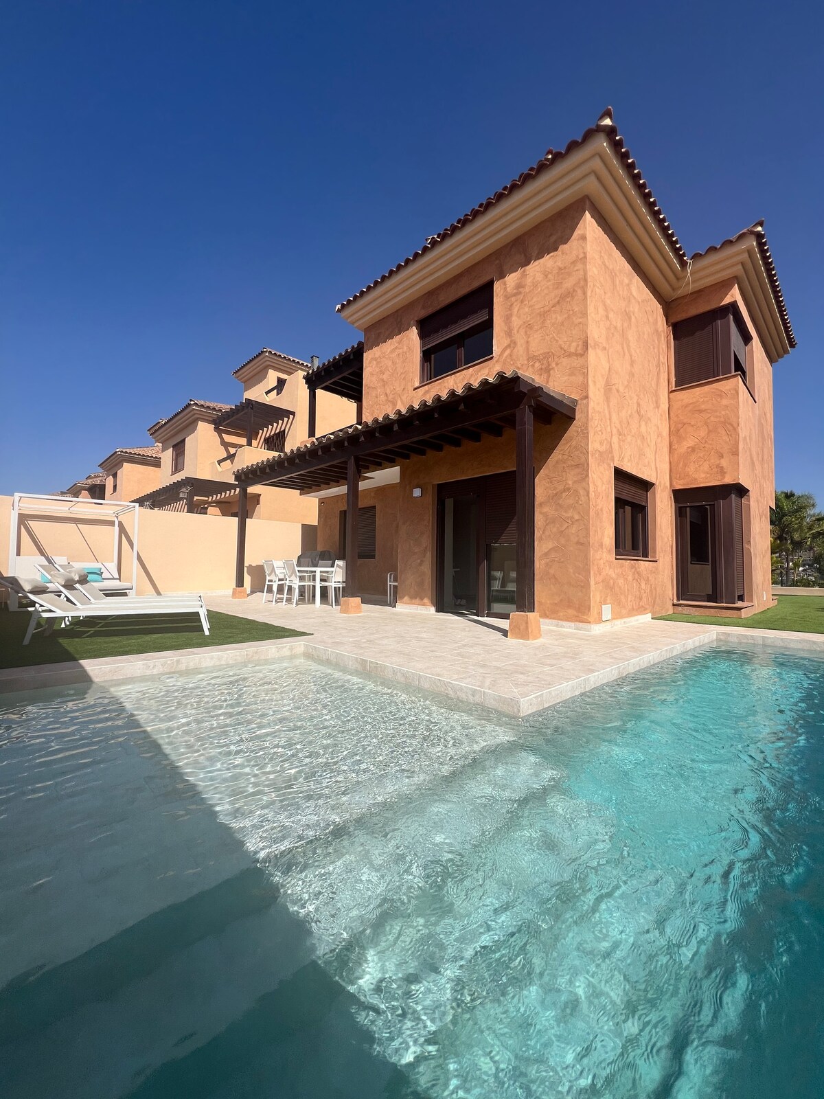 A modern two-story villa is set against a clear blue sky. The exterior features warm-toned walls and a shaded patio area. A private pool with clear water reflects the building's architectural lines, enhancing the outdoor space for relaxation.