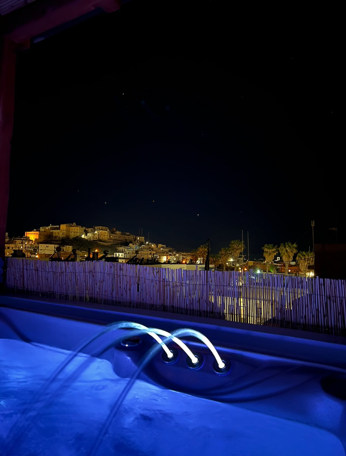 A view from the large balcony showcases a vibrant night scene, with illuminated buildings on a hillside. The calming blue light from the jacuzzi contrasts with the dark sky, enhancing the relaxed ambiance of the outdoor space.