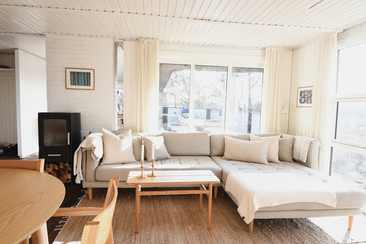 A modern living area features a spacious sectional sofa adorned with soft pillows, offering a comfortable seating option. Large windows allow natural light to fill the space, complementing the neutral color scheme. A wooden table is set nearby, enhancing the room's functionality.