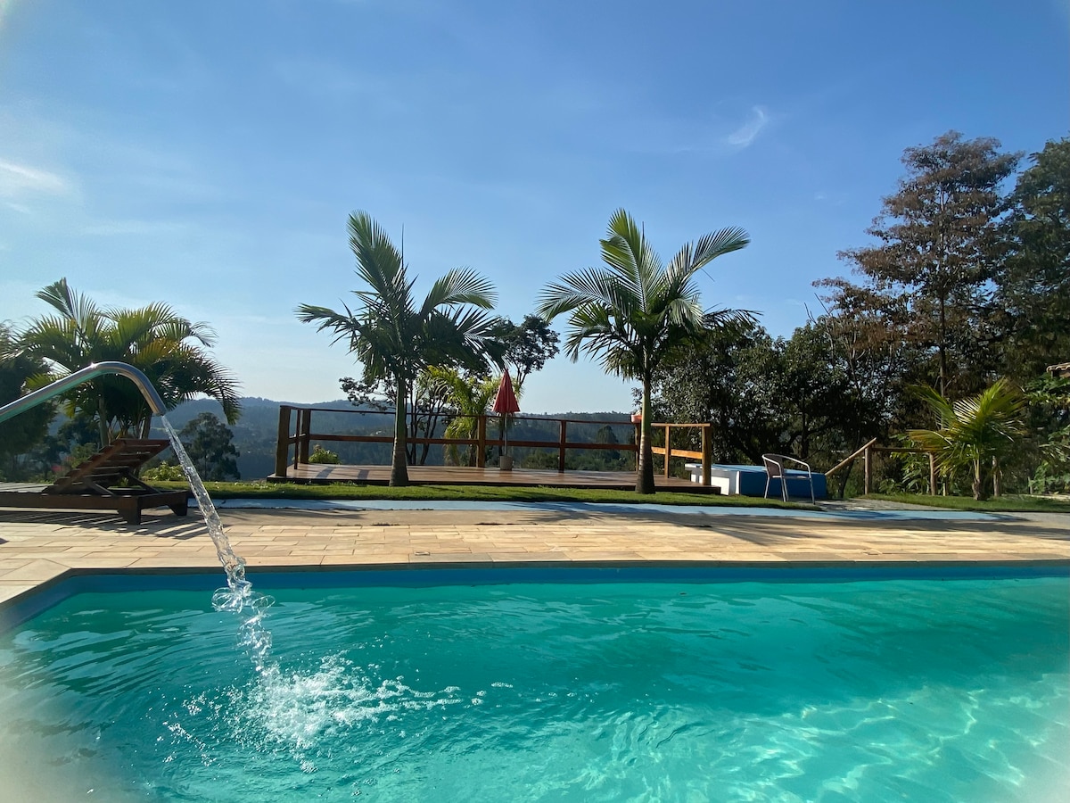 A serene swimming pool is highlighted in the foreground with a gentle waterfall feature. Lush palm trees and green hills create a natural backdrop, while wooden decking provides space for relaxation. Lounge chairs are positioned for enjoying the view under clear blue skies.