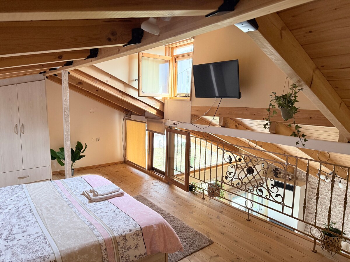 An airy loft space features a spacious bed adorned with soft linens and a light blanket. Natural light floods through multiple windows, illuminating the wooden beams and floor. A flat-screen TV is mounted on the wall, and potted plants add a touch of greenery.