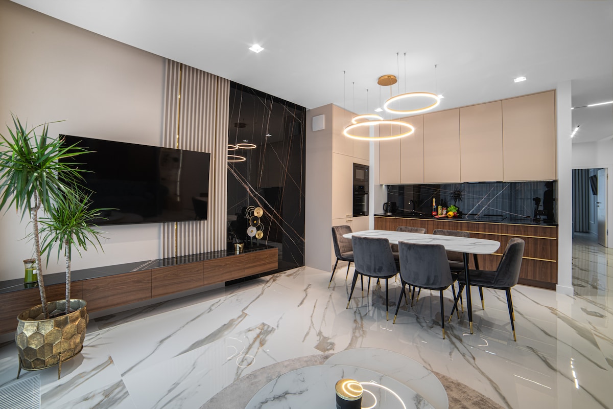 An open-plan living area features a modern kitchen with a sleek countertop and dining space, accommodating six upholstered chairs around a round table. A large wall-mounted television is positioned above a stylish wooden console, while a decorative plant adds a touch of greenery to the space.