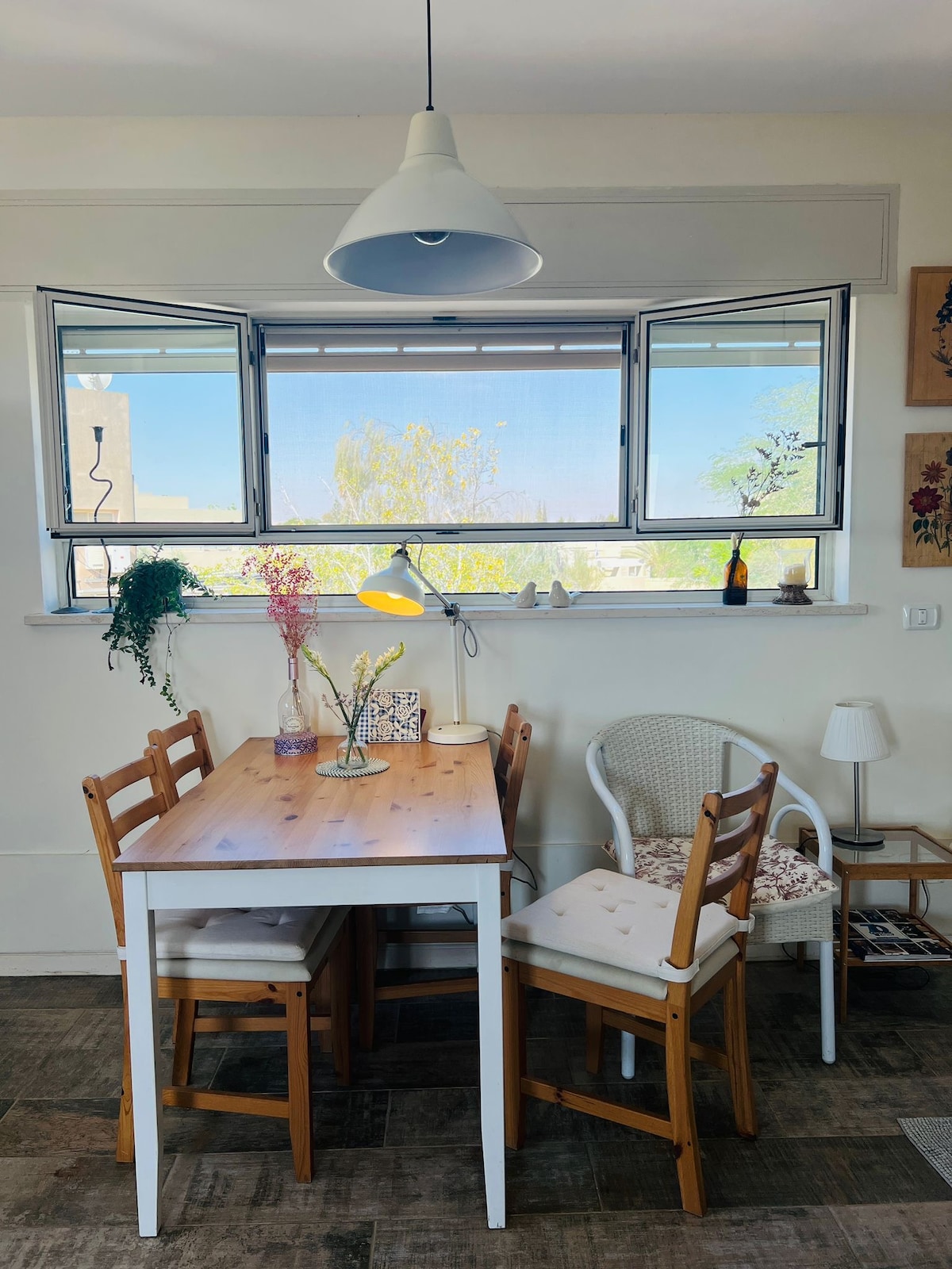 A dining area is featured with a wooden table and four chairs, two of which are white wicker. Natural light fills the space through large, open windows, accentuated by a pendant light overhead. Decorative plants and a small vase add a touch of greenery.