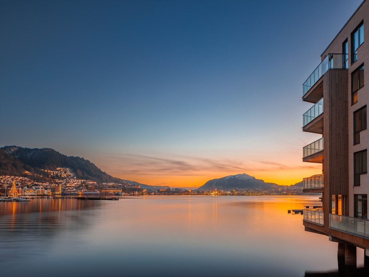 A serene view of the water at sunset reflects shades of orange and blue across the calm surface. The mountains are visible in the distance, while modern buildings with glass balconies line the edge of the water, creating a harmonious blend with the natural landscape.