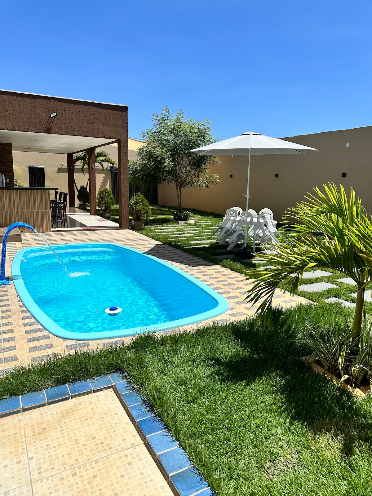 A refreshing swimming pool is surrounded by manicured grass and decorative stones, with a comfortable umbrella providing shade over white lounge chairs. Nearby, a cozy outdoor seating area is nestled under a roof, enhancing the inviting and spacious ambiance of the garden.