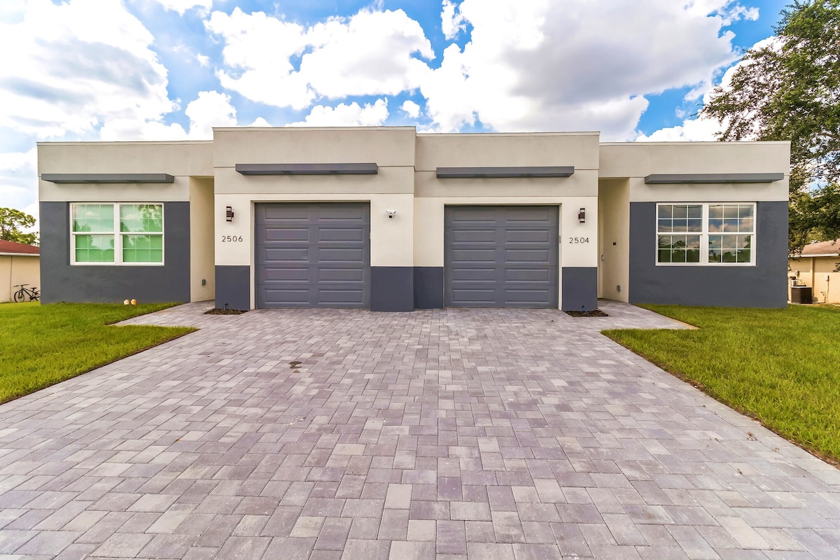 A modern duplex with a symmetrical facade is set against a partly cloudy sky. The structure features two separate garages with matching doors, framed by large windows on either side. A well-maintained paver driveway stretches forward, bordered by green grass.