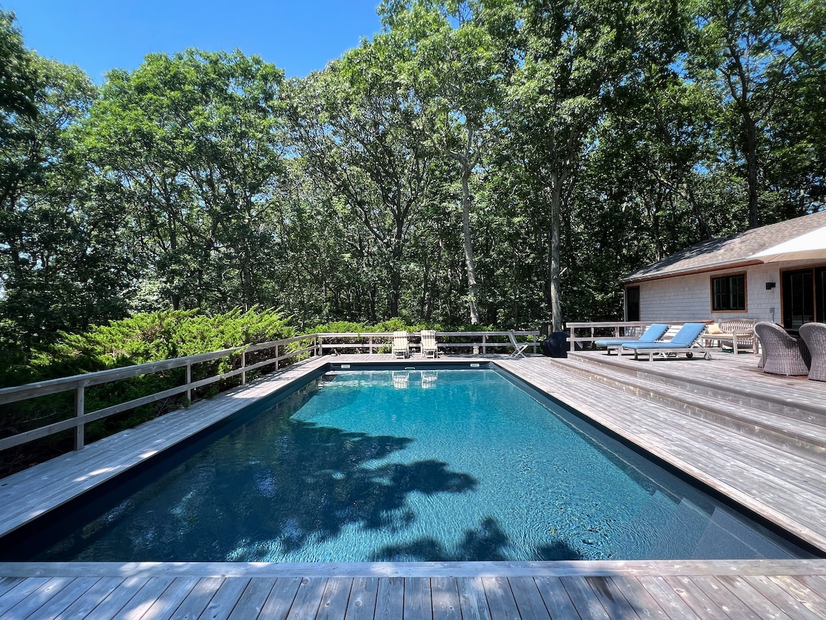 A large swimming pool is set within a wooden deck, surrounded by lush greenery. The pool's clear blue water reflects the sunlight, while several lounge chairs are placed around the deck, providing ample space for relaxation. The tranquil setting enhances a sense of privacy.
