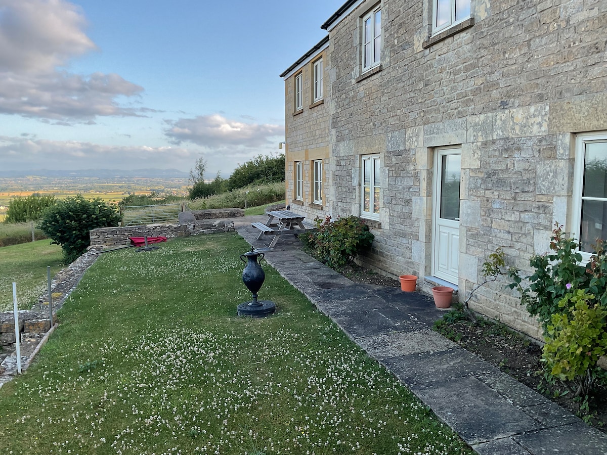 The exterior of a stone farmhouse is viewed from the garden, showcasing a well-kept lawn dotted with white flowers. A patio area features a table and chairs, with potted plants placed near the entrance. Scenic views of rolling hills are visible in the background.