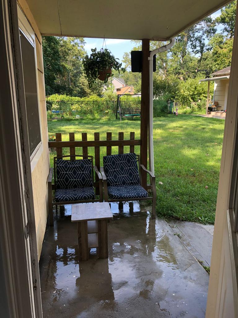 An outdoor seating area features two patterned chairs positioned beside a small wooden table. The concrete floor reflects sunlight, with lush greenery visible in the background. A gated area and a glimpse of additional outdoor space can be seen, providing a serene setting.