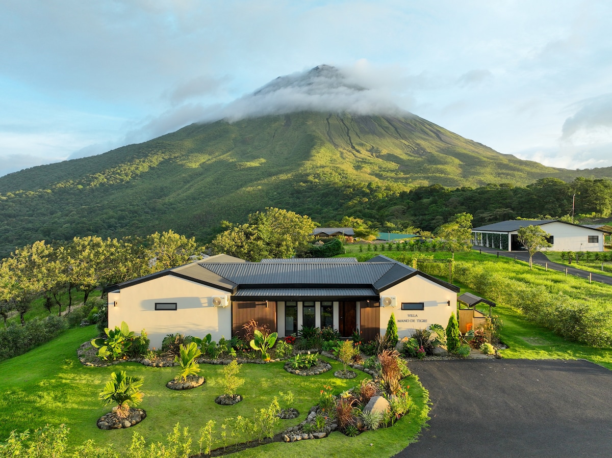 A contemporary villa is set against the backdrop of Arenal Volcano, featuring a well-manicured garden with various tropical plants. The structure's modern design includes a combination of horizontal and vertical lines, while a spacious driveway leads up to the main entrance.