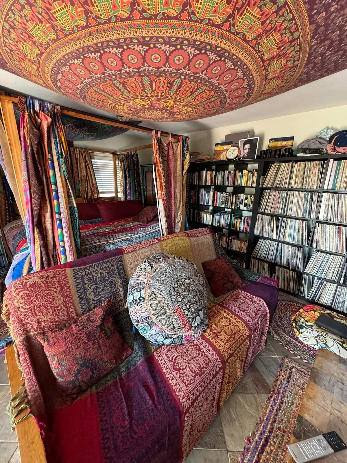 A vibrant seating area features a richly colored couch adorned with decorative pillows and a patterned throw. Walls are draped in colorful fabric, and a large tapestry hangs from the ceiling. A collection of vinyl records is neatly organized on nearby shelves.