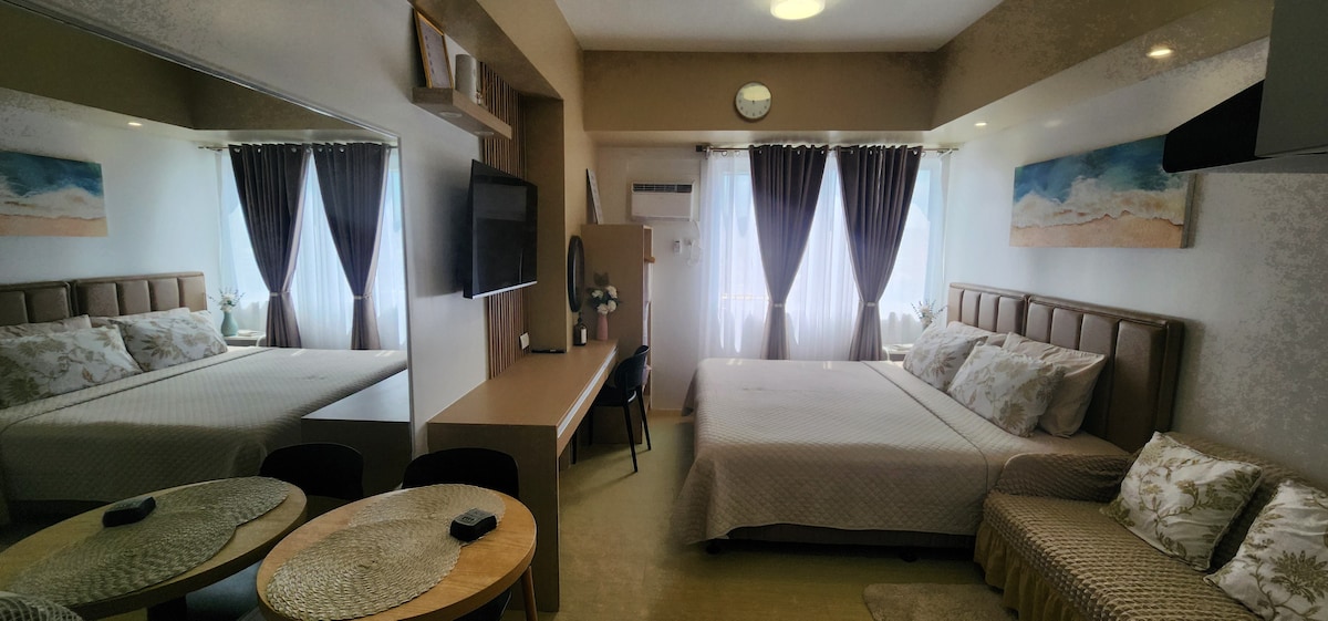 The room features a spacious king-size bed with decorative pillows and soft linens. A wide working table is positioned near a wall-mounted television. Natural light fills the space through two large windows, alongside a comfortable 2-seater couch and dining area, enhancing the inviting ambiance.