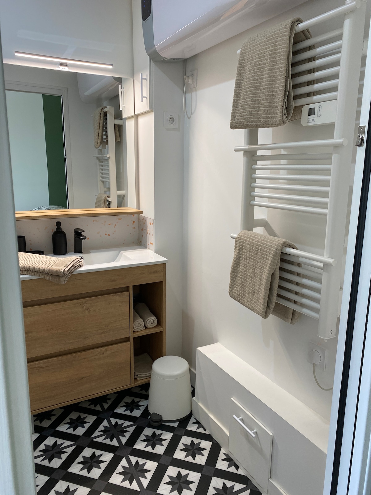 The bathroom features a modern sink area with wooden cabinetry, accompanied by neatly arranged toiletries. A towel rack holds folded towels, and a white waste bin is positioned on the floor. The patterned floor tiles add an interesting visual element to the space.
