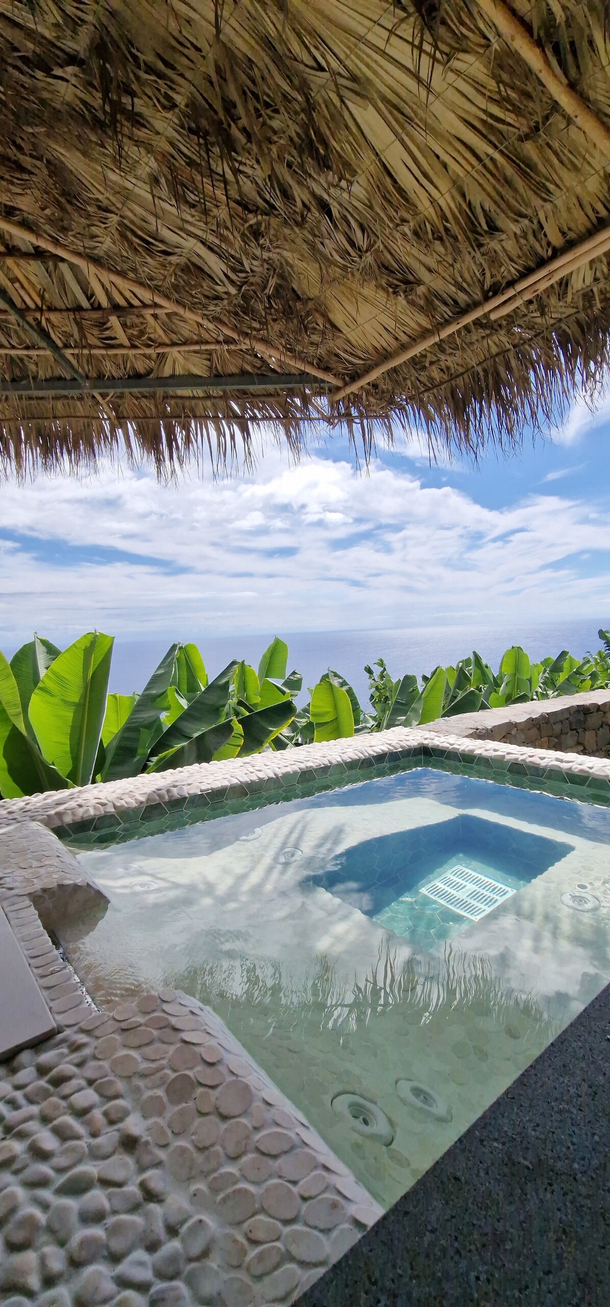 A private hot tub is set within a stone border, surrounded by lush green foliage. A thatched roof provides shade, while the distant view of the Atlantic Ocean and cloudy sky is visible in the background.