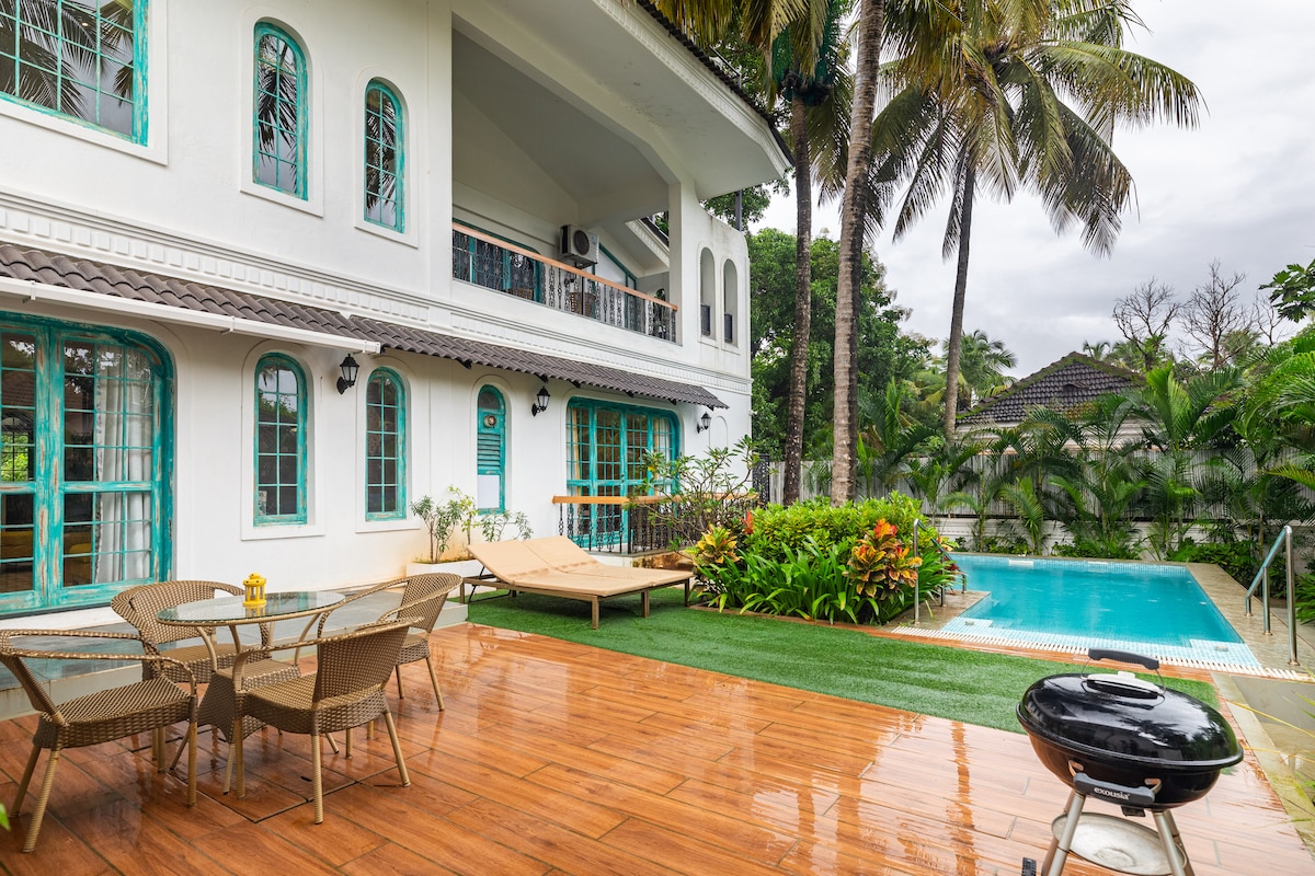 An outdoor patio features a dining table with wicker chairs and sun loungers, complemented by a barbecue grill. Lush greenery surrounds the area, with a turquoise swimming pool visible nearby, and a spacious villa stands in the background beneath a cloudy sky.