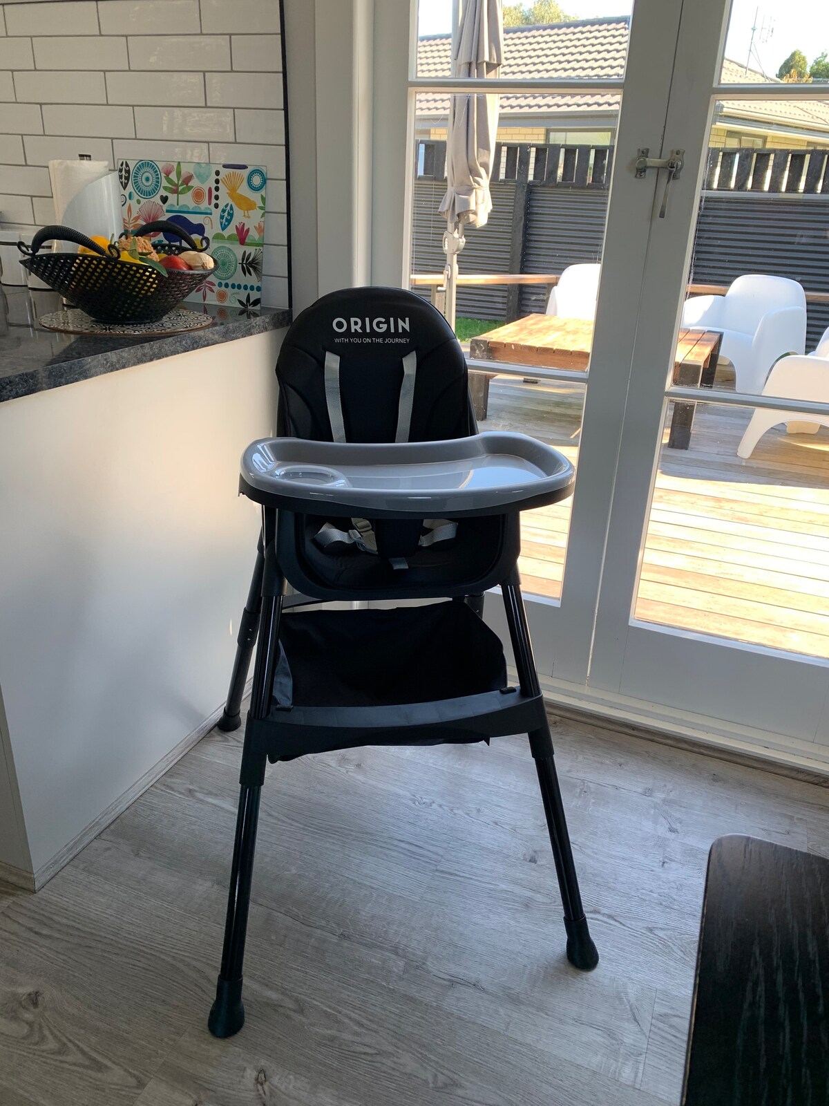 A modern high chair is positioned beside a countertop, featuring a sleek black design with a removable tray. Large glass doors allow natural light to illuminate the area, and a spacious deck with white outdoor furniture is visible in the background.