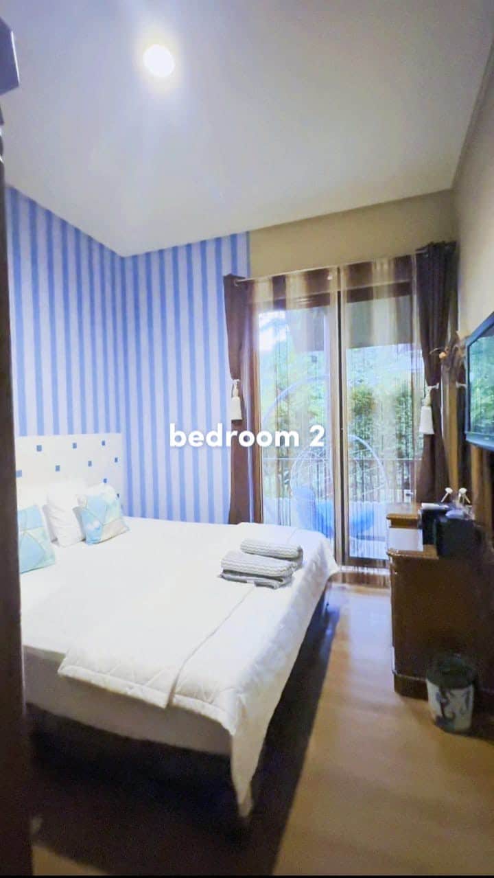 A bright bedroom is characterized by blue-striped walls and a spacious bed with neatly arranged towels. Natural light filters through glass doors leading to a balcony, enhancing the room’s cozy atmosphere. A wooden nightstand and a flat-screen TV add functional elements.