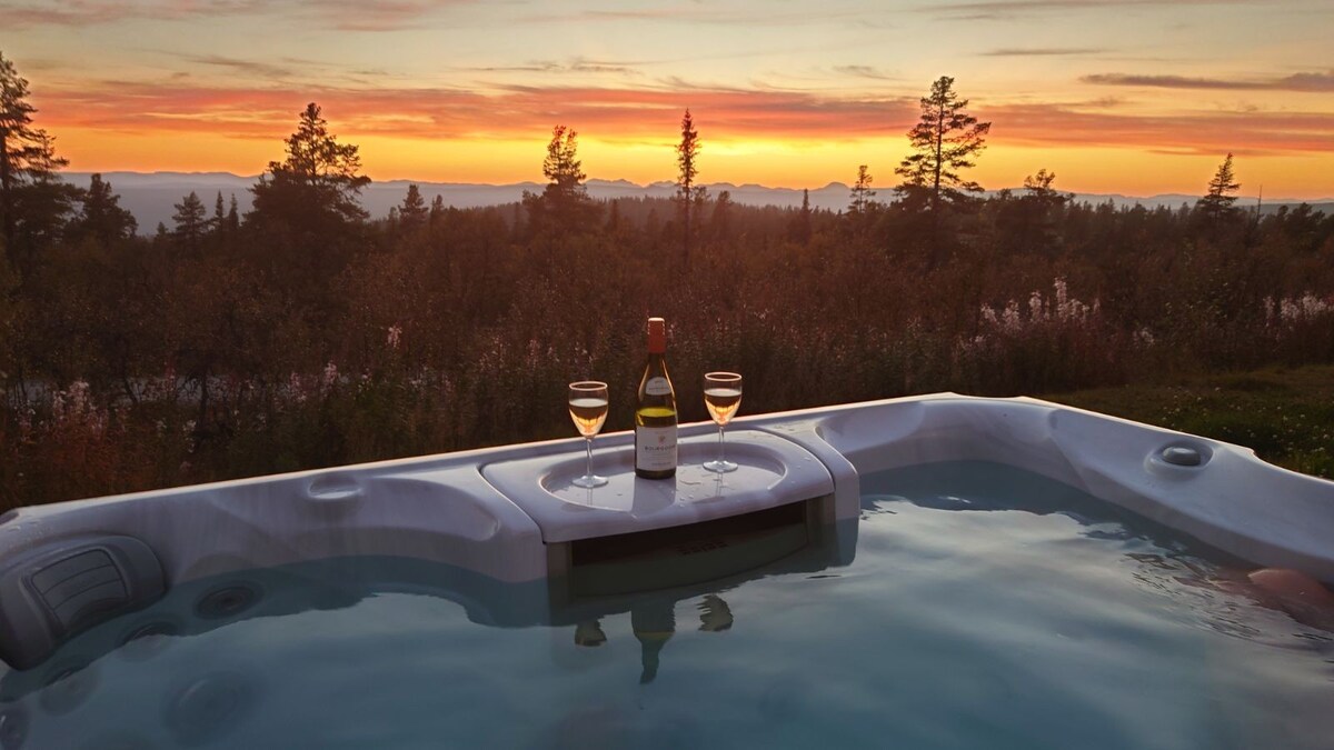 The outdoor Jacuzzi offers a scenic view of the sunset, surrounded by lush vegetation. A bottle of wine rests on the ledge alongside two glasses, inviting relaxation and enjoyment amidst nature.