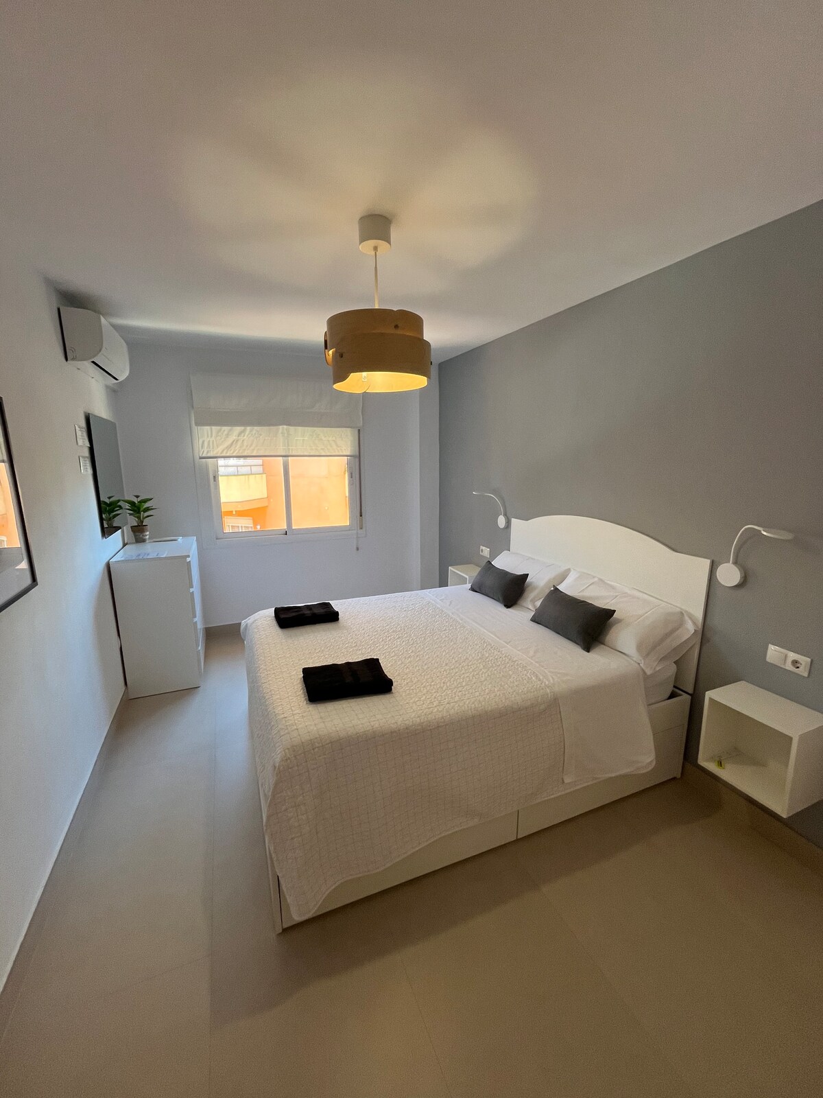 A bright bedroom is highlighted by soft natural light coming through a window. The room features a spacious bed with a white quilt and dark accent pillows, complemented by a bedside table and a dresser. A simple pendant light provides additional illumination.