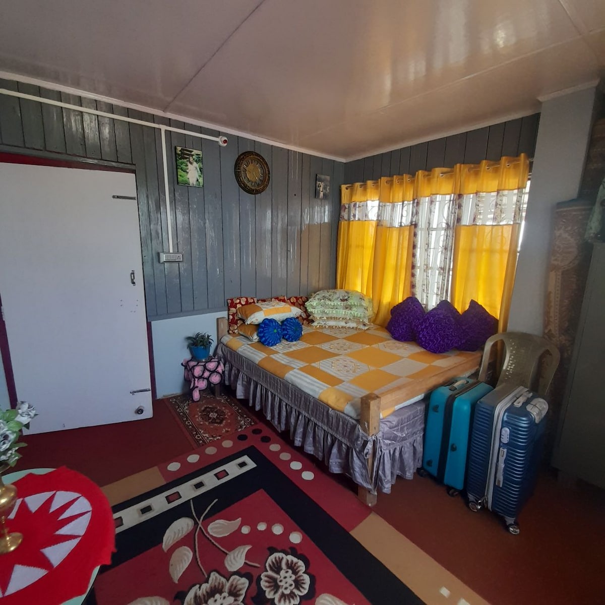 A seating area designed for relaxation is showcased with a spacious bed adorned with colorful cushions. Bright yellow curtains filter sunlight, while a patterned area rug adds texture. Two packed suitcases and a decorative table with floral patterns are also visible, contributing to the room's inviting character.