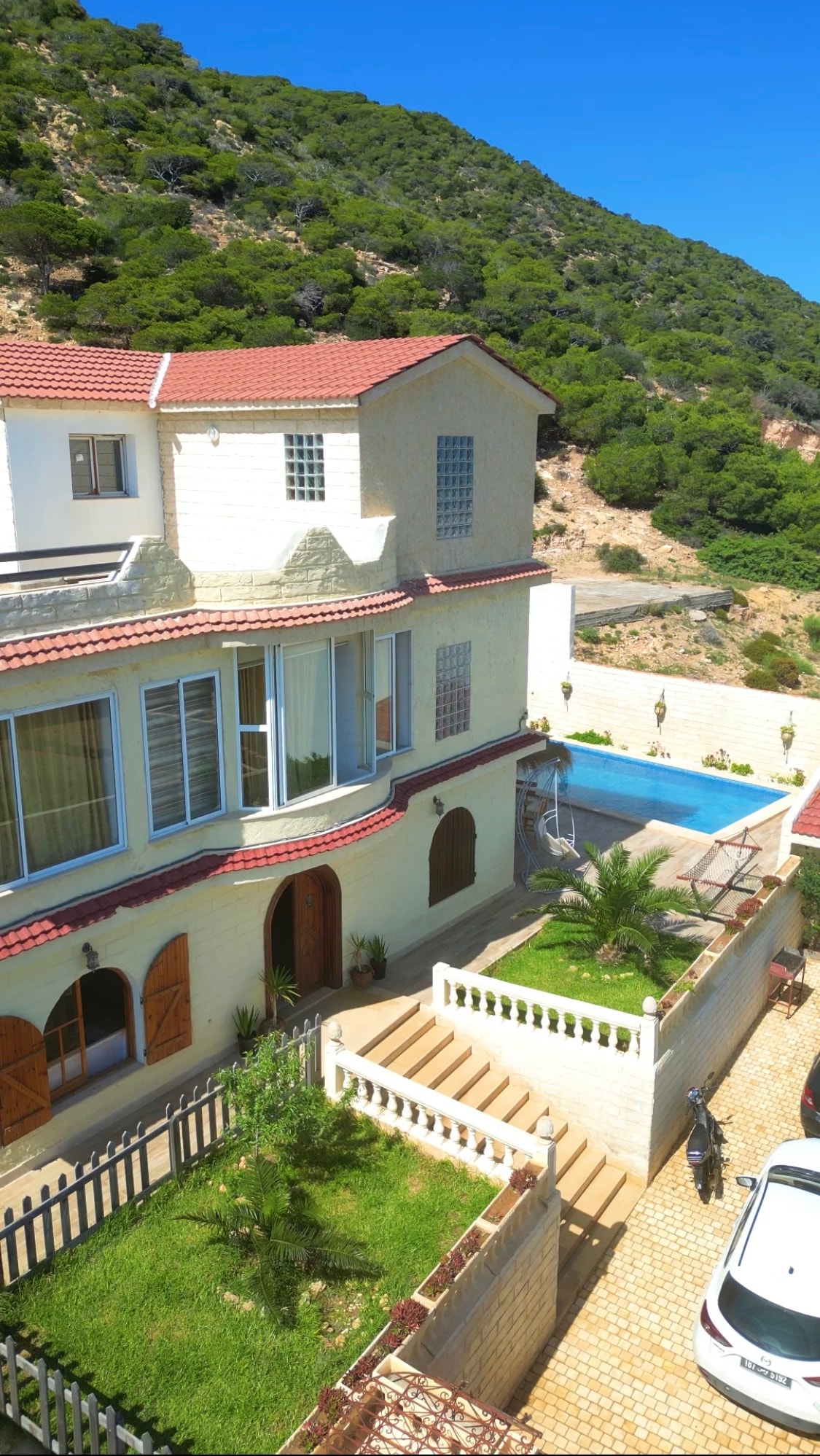 An elevated view of the elegant property reveals a charming exterior with light-colored walls and a red-tiled roof. The inviting pool is bordered by greenery, with steps leading down to the entryway. Surrounding mountains create a serene backdrop.