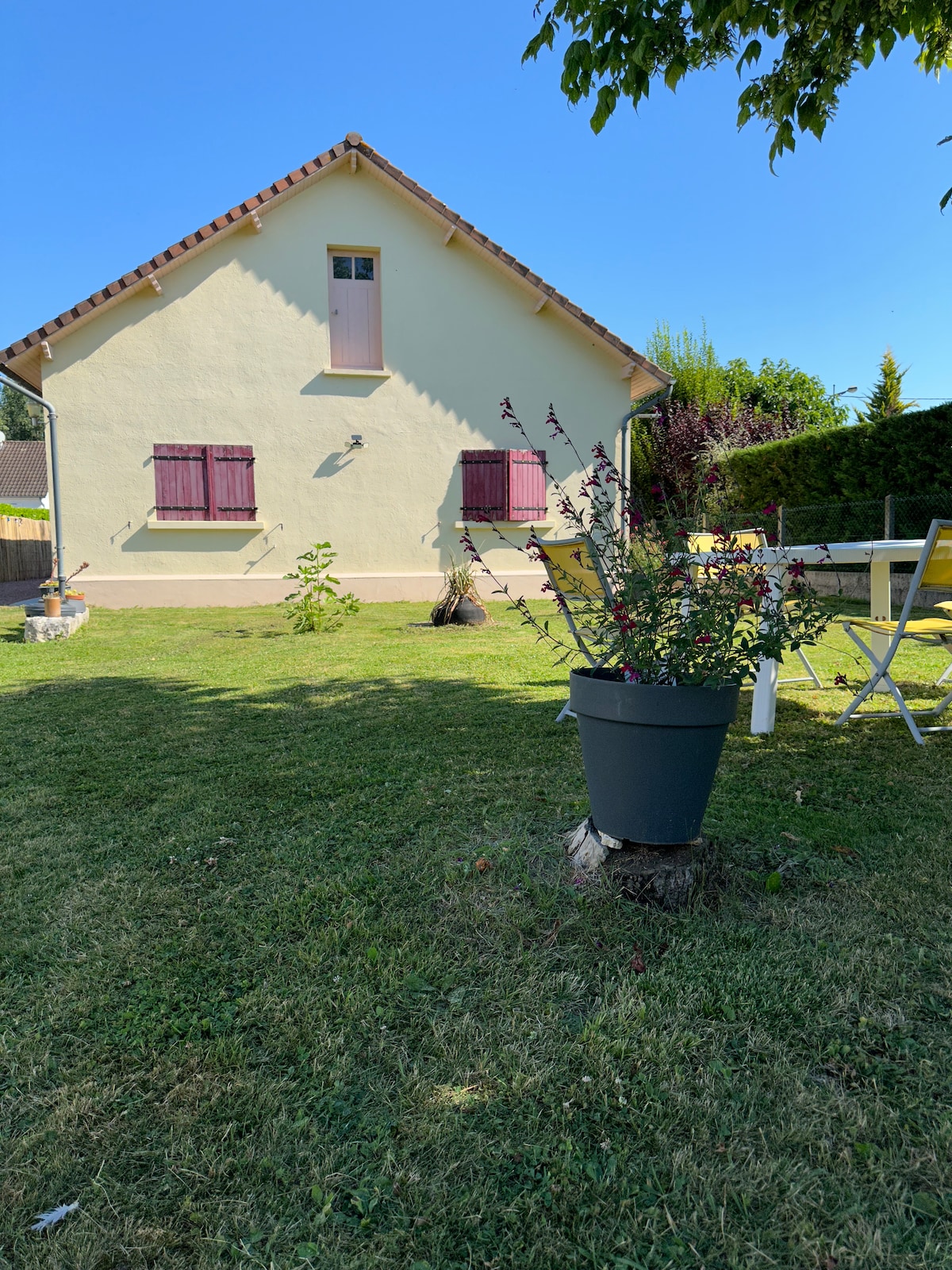 The image showcases a peaceful garden with a well-maintained lawn and flower arrangements. A light-colored house with purple shutters is visible in the background. A table with chairs is placed outdoors, creating a welcoming space for lounging or dining.