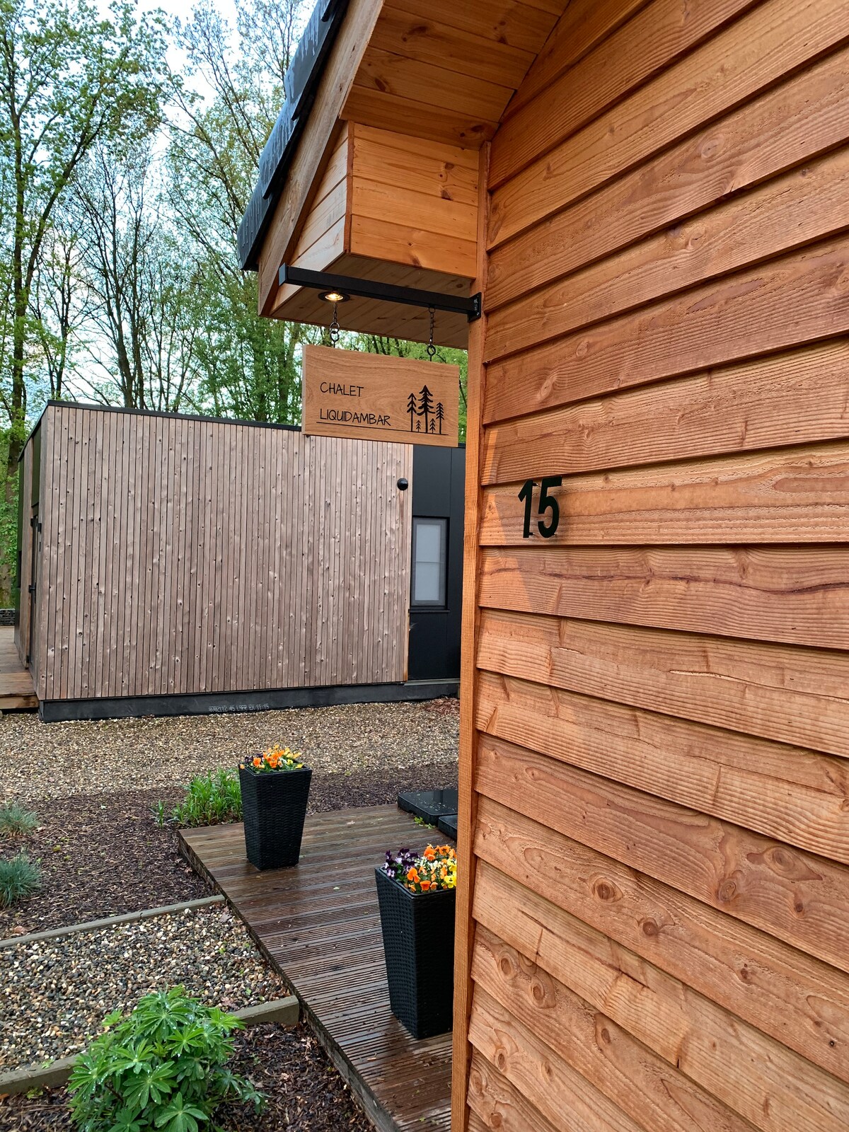 The exterior of a wooden chalet is showcased, featuring natural wood siding and a clear, numbered sign. Nearby, a modern structure is visible, surrounded by landscaped stone pathways and potted flowers, creating a serene setting in a wooded area.