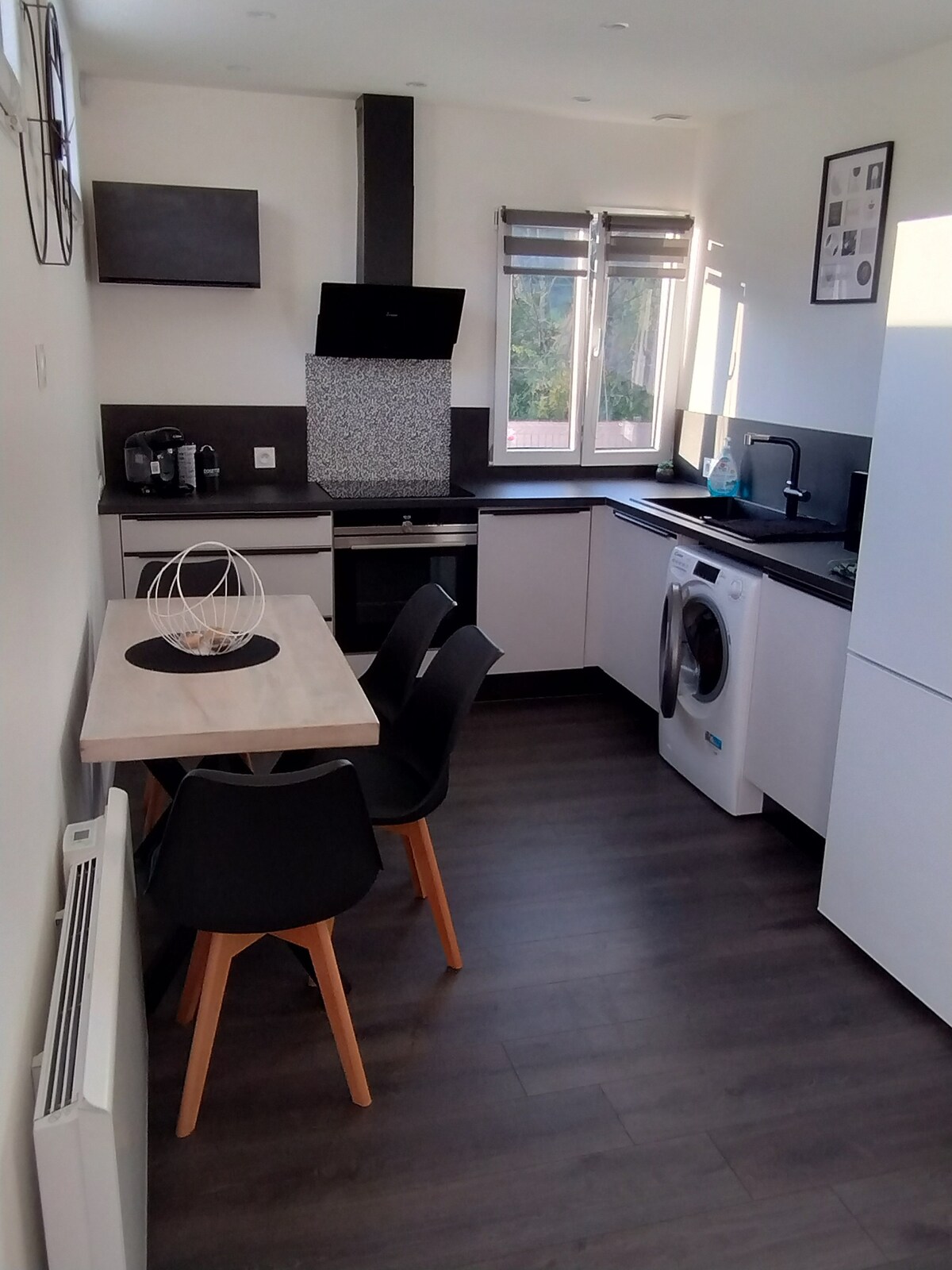 The kitchen area features modern, sleek cabinetry and a compact dining table with four chairs. An equipped cooking space includes a stove, oven, and microwave. A laundry machine is discreetly integrated into the design. Natural light enters through the windows, enhancing the bright atmosphere.