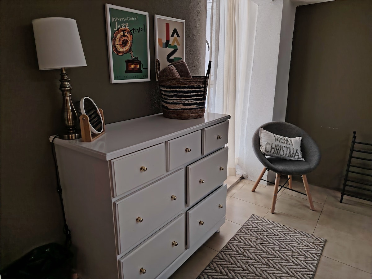 A well-organized dresser with multiple drawers is positioned against a grey wall. A modern lamp stands beside a woven basket. Art pieces are displayed on the wall, while a comfortable chair adds a cozy touch to the space. Natural light filters in through a sheer curtain.