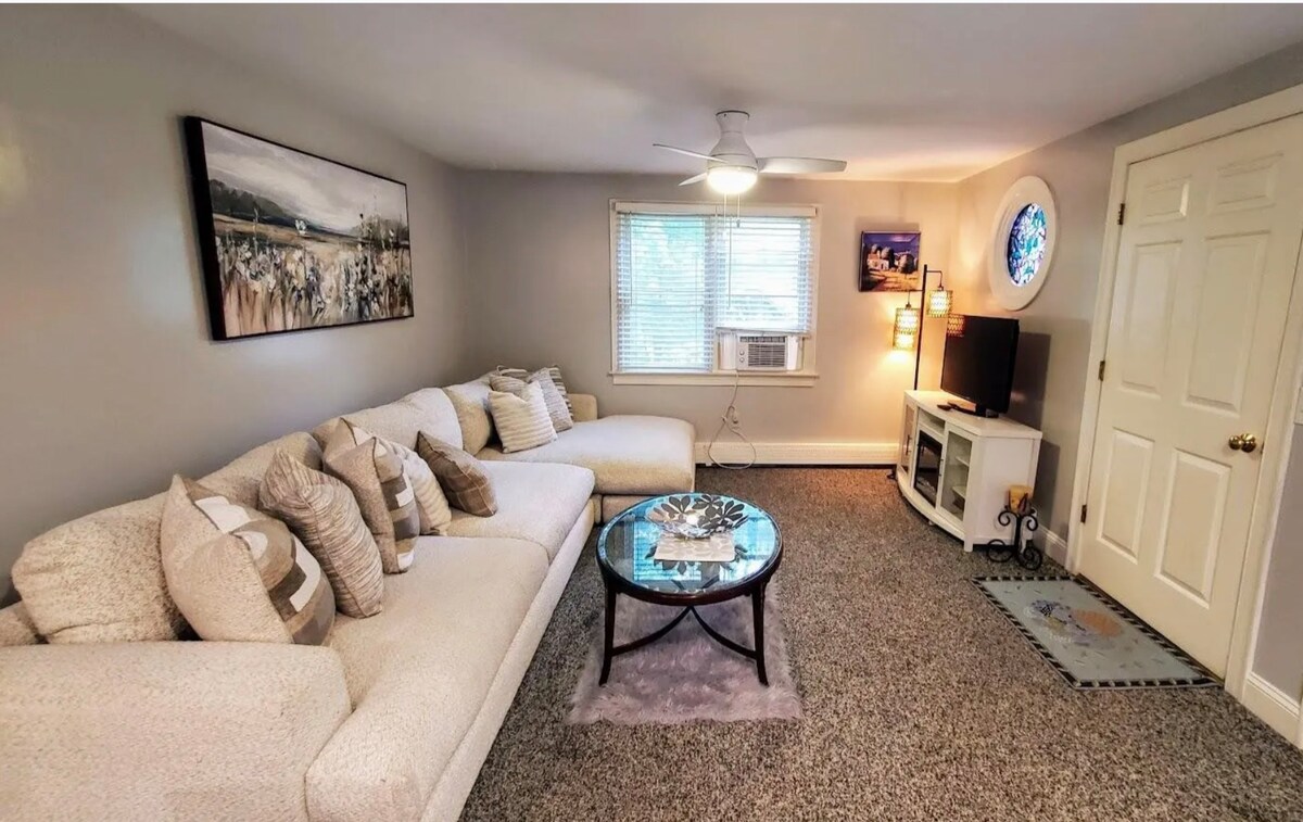 A spacious living area features a large light-colored sectional sofa adorned with patterned throw pillows. A circular glass coffee table is centrally positioned. The room is brightened by natural light through a window, with a wall-mounted television and an air conditioning unit visible.