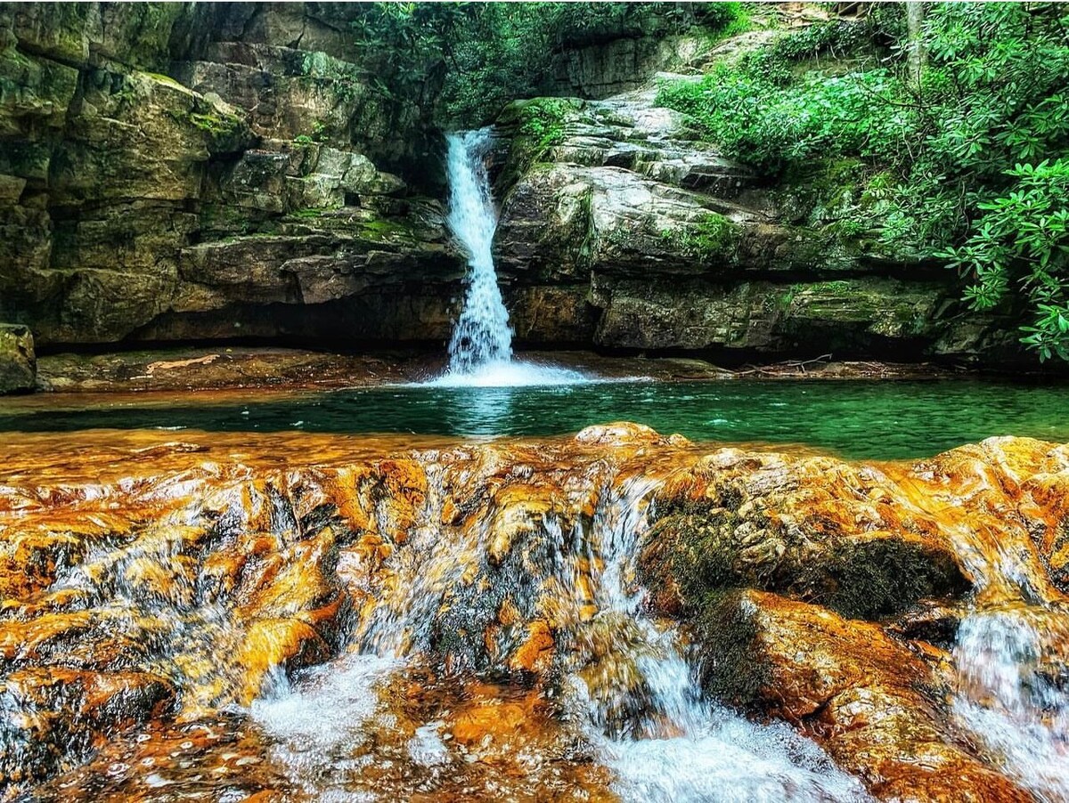 A cascading waterfall flows over smooth, layered rocks into a clear pool, surrounded by lush greenery. The vibrant orange and brown hues of the rocks contrast with the cool blue-green water, creating a serene natural setting.
